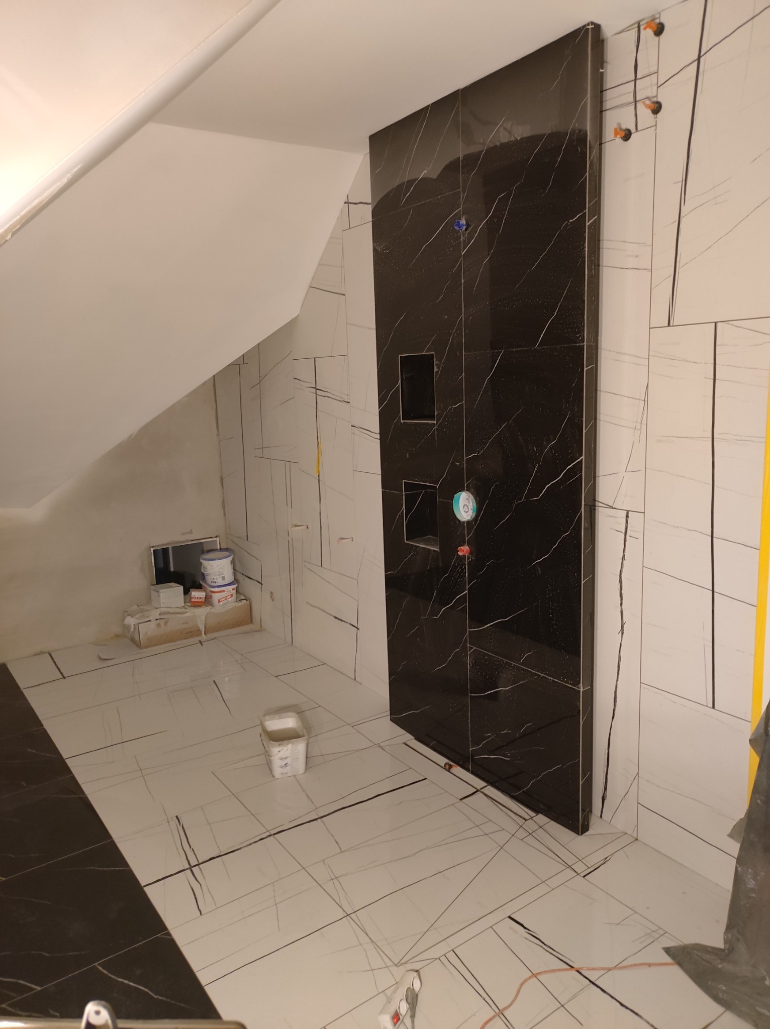 Łazienka w trakcie remontu: czarne płytki imitujące marmur na ścianie prysznicowej z wnękami, białe płytki podłogowe i ścienne z szarymi żyłami, widoczne materiały budowlane i narzędzia, poddasze...