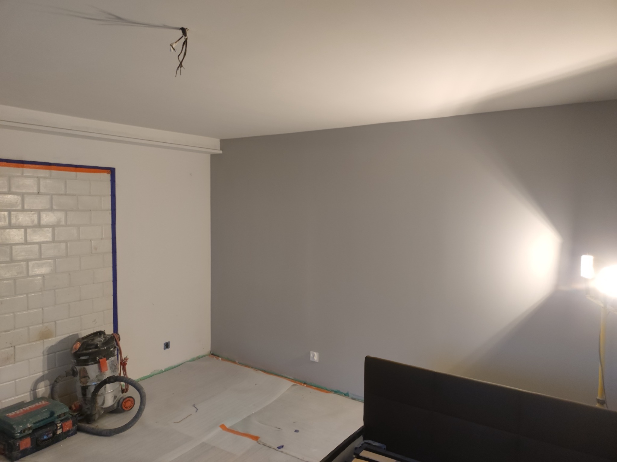 Pokój w trakcie malowania na szaro, zabezpieczona folią podłoga, widoczny odkurzacz przemysłowy i narzędzia, fragment łóżka w prawym dolnym rogu.