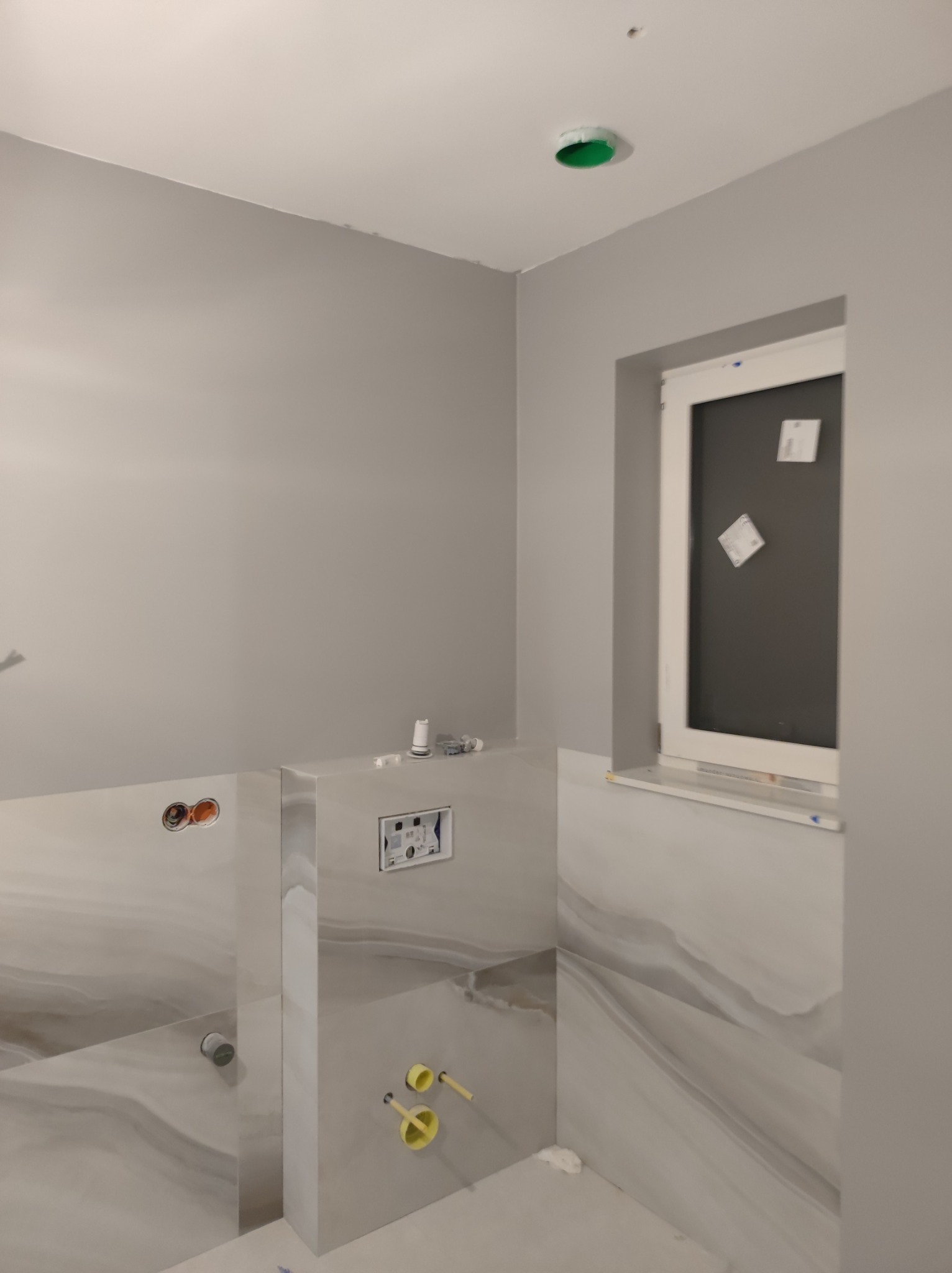 Szara łazienka w trakcie remontu: widoczne przyłącza wodne, zabudowa stelaża WC z płytkami imitującymi marmur, okno z przyklejonymi kartkami, oraz zielony element instalacji na suficie.