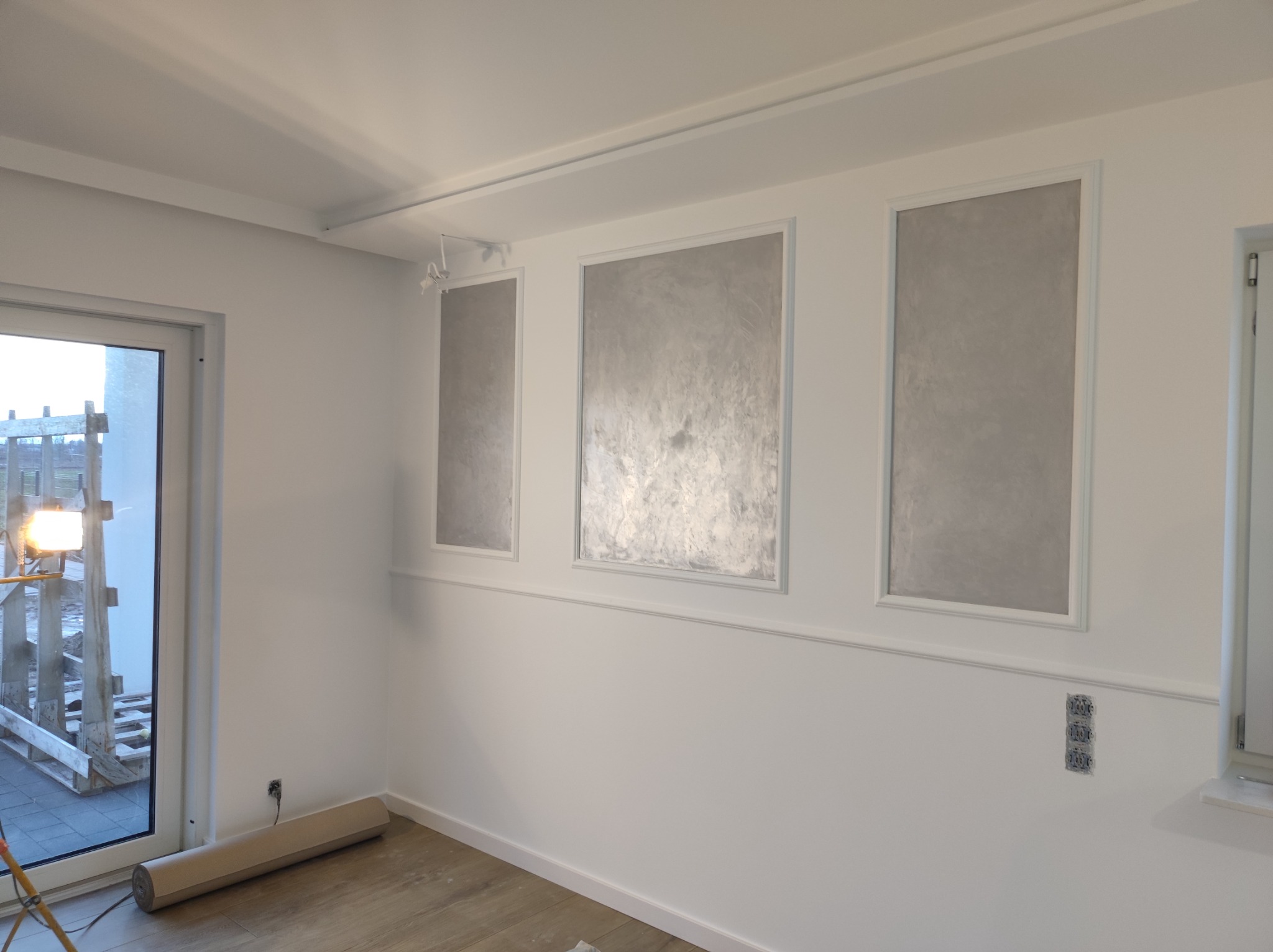 Pokój po remoncie z trzema dekoracyjnymi panelami na ścianie, widoczna rolka zabezpieczająca podłogę oraz lampa budowlana przy oknie.