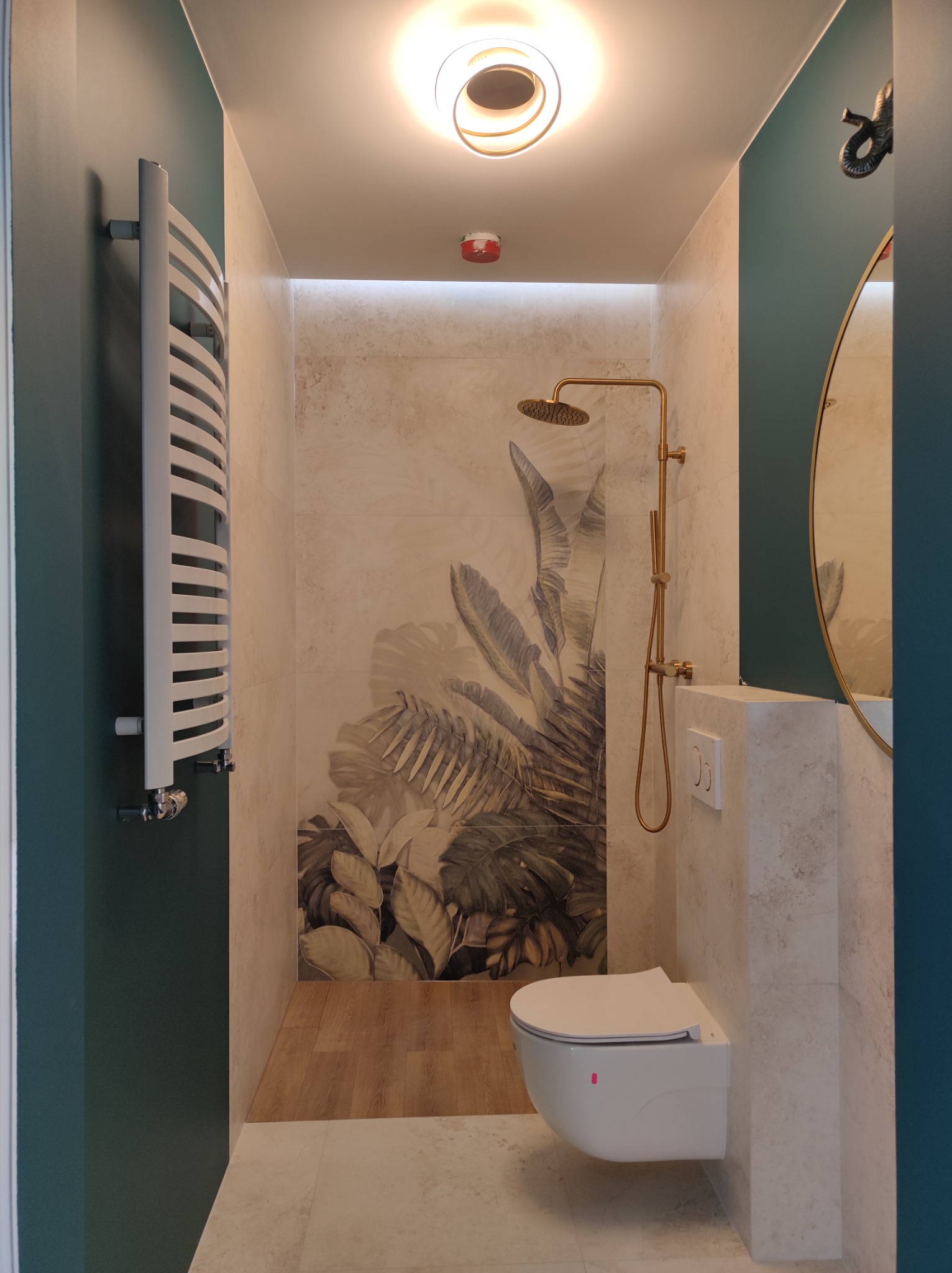 Nowoczesna łazienka z motywem tropikalnych liści na ścianie, złota armatura prysznicowa, podwieszana toaleta i grzejnik drabinkowy.