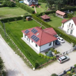 Dom jednorodzinny z panelami słonecznymi na czerwonym dachu, otoczony zielonym trawnikiem i tujami, widziany z lotu ptaka w słoneczny dzień.