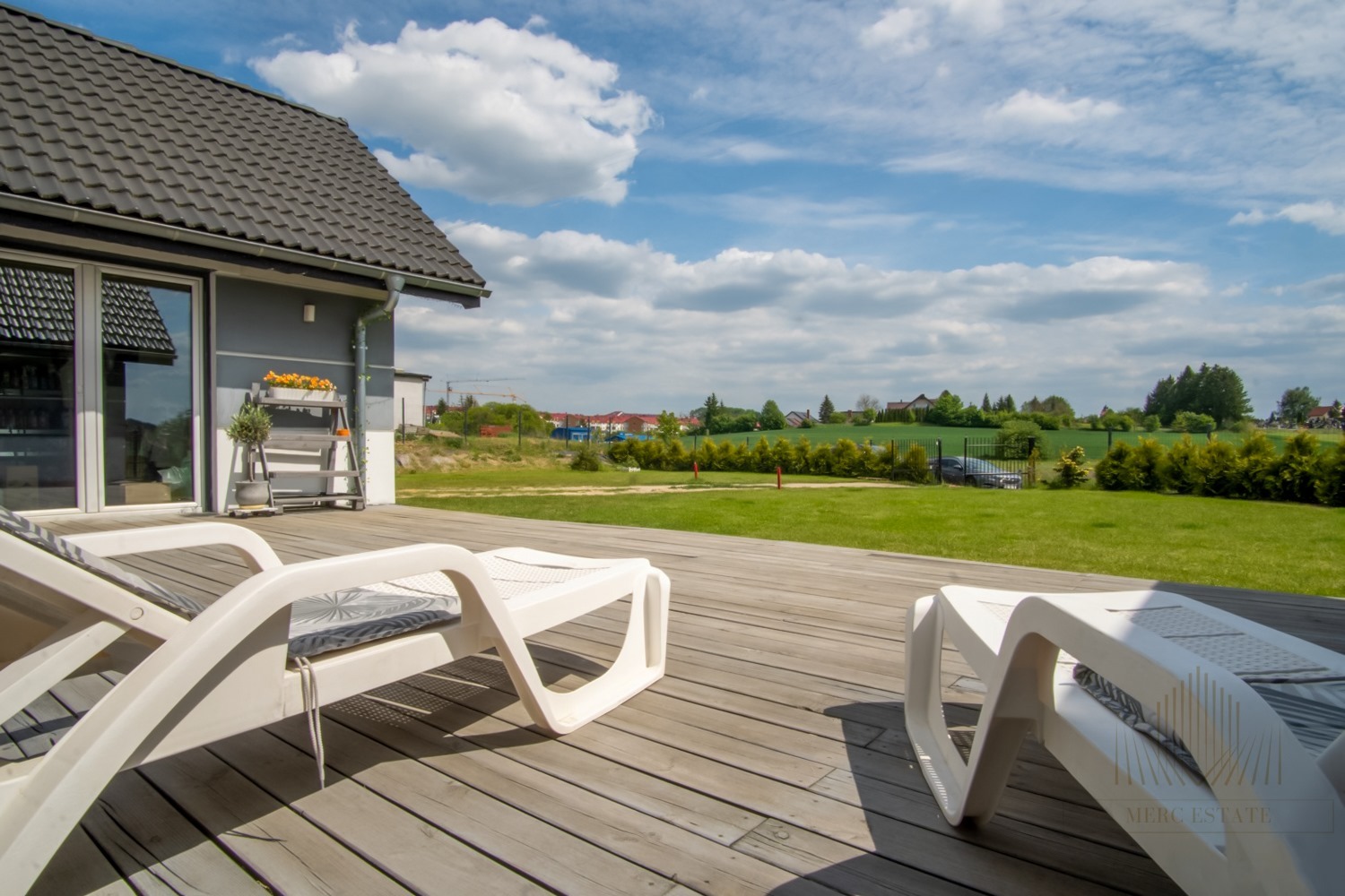 Nowoczesny dom z tarasem i leżakami w słoneczny dzień. Widok na zadbany trawnik i okoliczną zabudowę. Jasne kolory i przestrzeń.