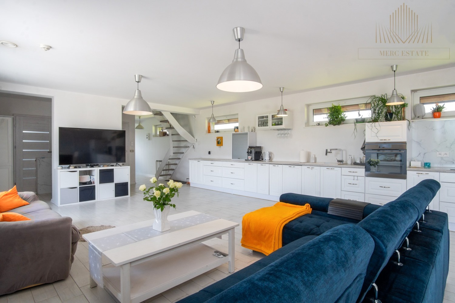 Jasne wnętrze domu z otwartą kuchnią, schodami i salonem. Wystrój w bieli, szarości i akcentach kolorystycznych. Widoczny narożnik i kwiaty.