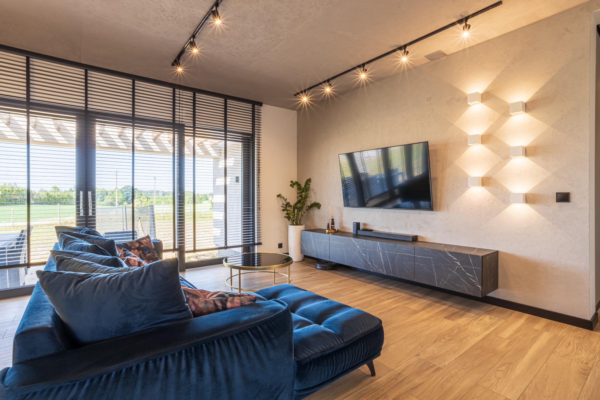 Nowoczesny salon: granatowa sofa, drewniana podłoga, TV na ścianie, designerskie oświetlenie i widok na zieleń przez okno z żaluzjami.