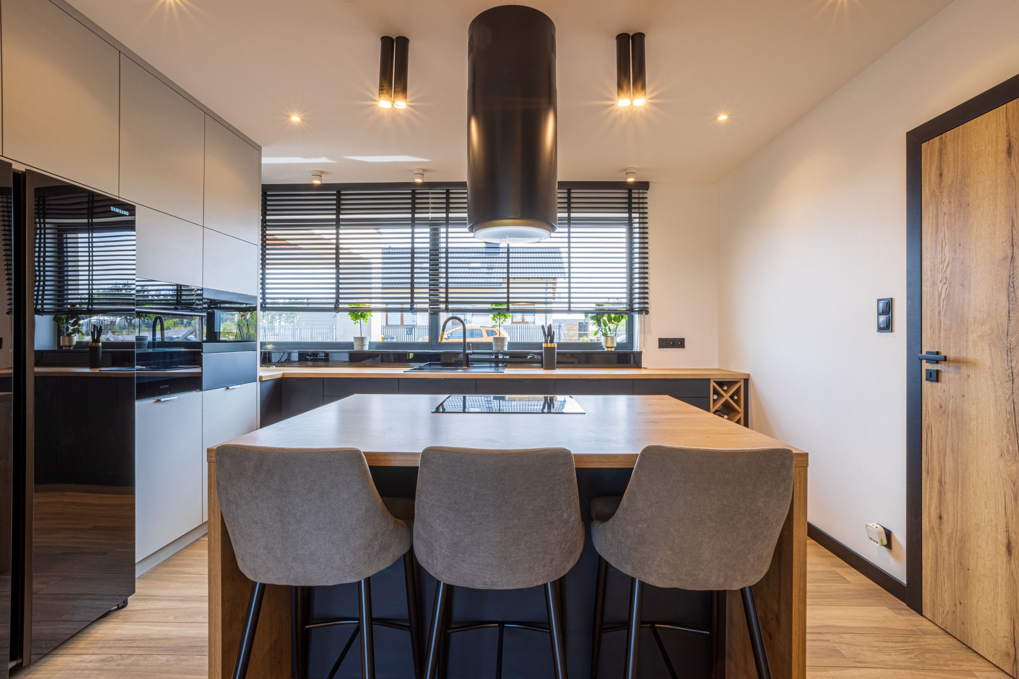 Nowoczesna kuchnia z wyspą i trzema krzesłami barowymi. Czarny okap cylindryczny, drewniane blaty i podłoga, duże okno z żaluzjami, lodówka z czarnym frontem.