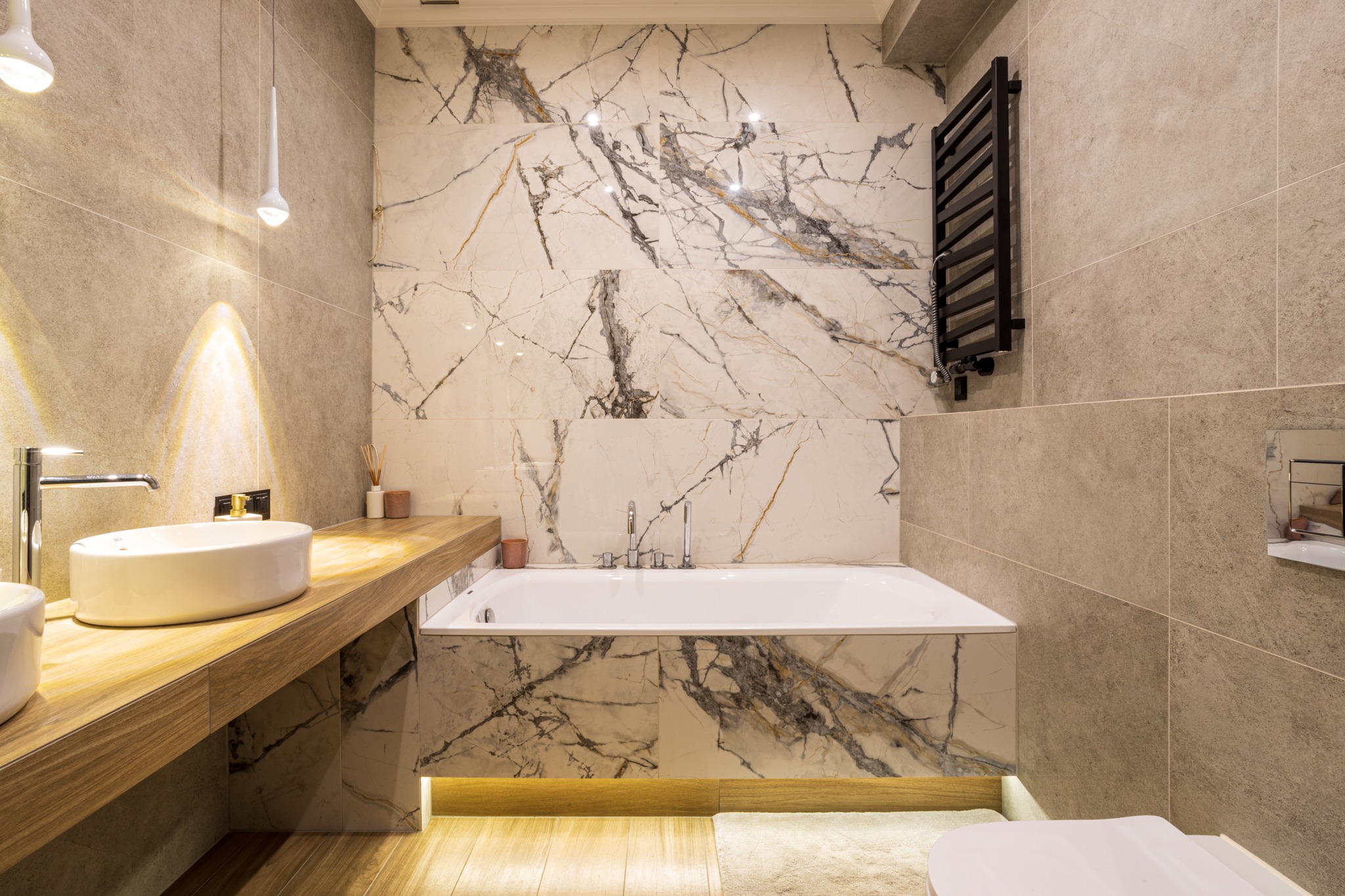 Elegancka łazienka z wanną obudowaną marmurem, drewnianym blatem pod umywalką i grzejnikiem drabinkowym. Nowoczesny design i stonowana kolorystyka.