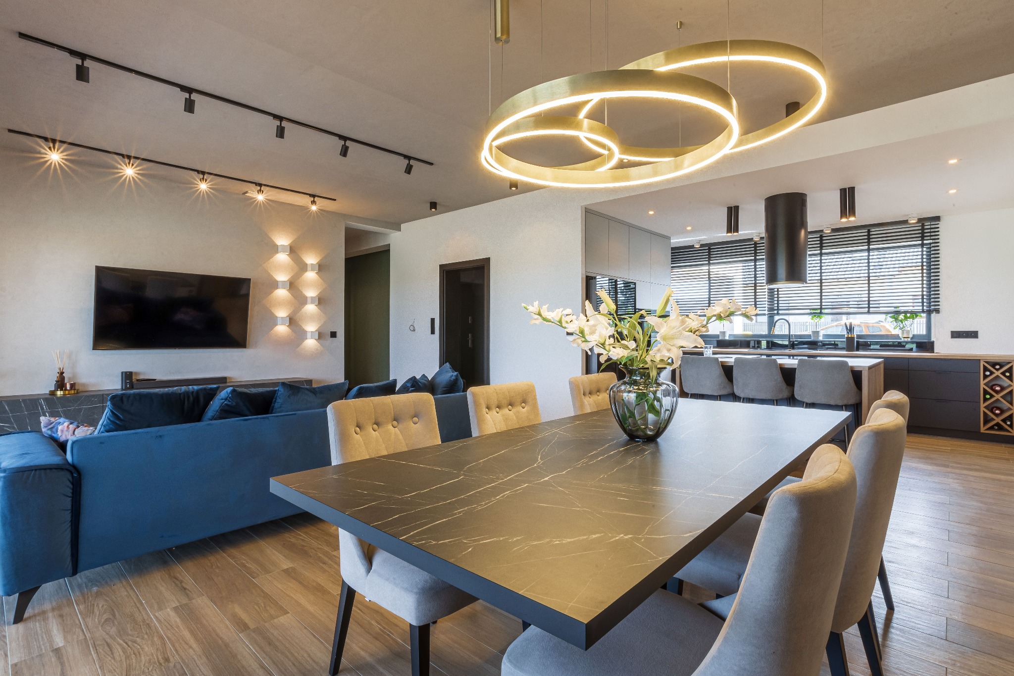 Eleganckie wnętrze: stół z marmurowym blatem, kwiaty w wazonie, niebieska sofa, otwarta kuchnia. Nowoczesne oświetlenie podkreśla styl.