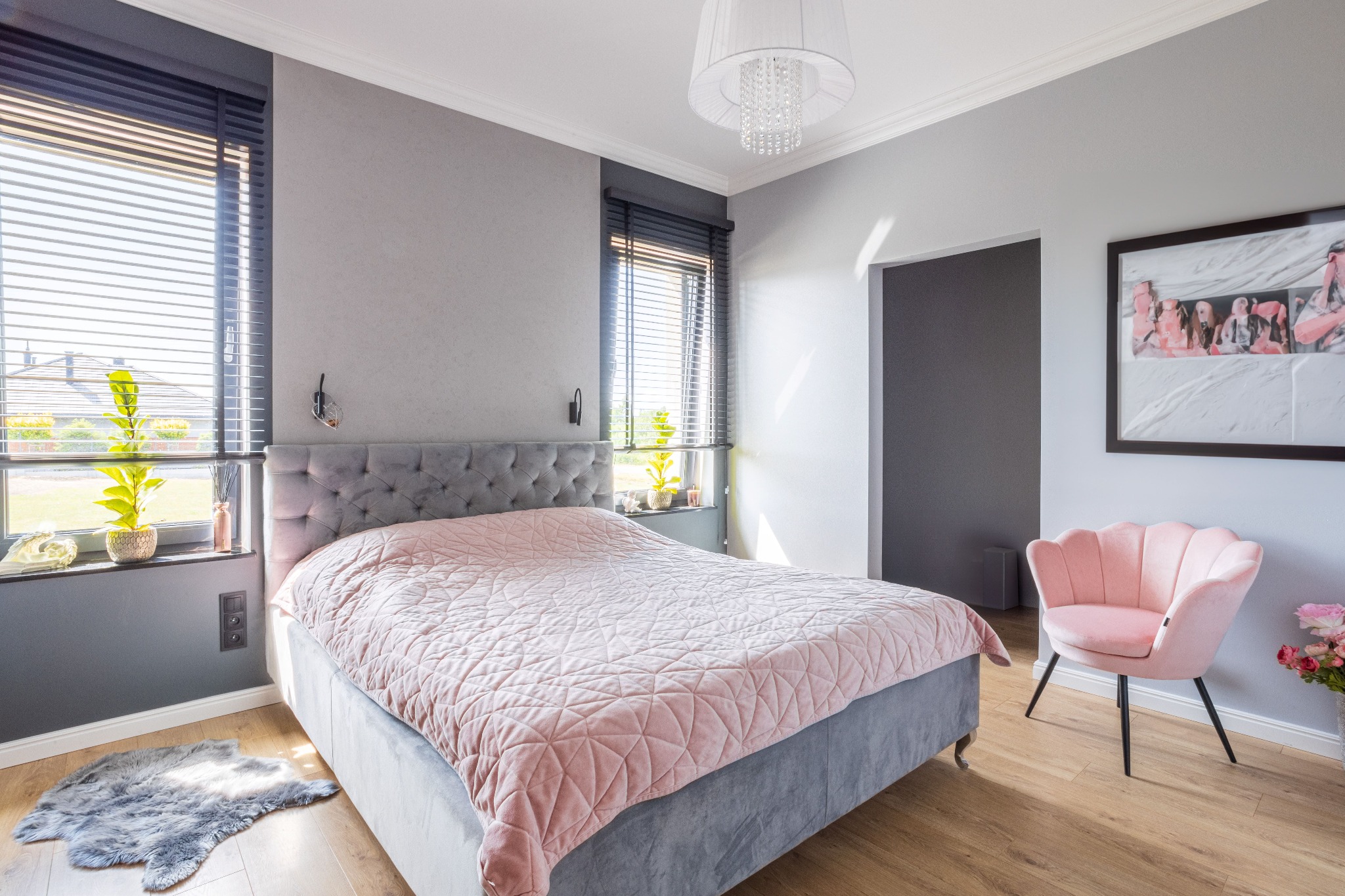Elegancka sypialnia z szarą tapetą i łóżkiem, różowa narzuta, drewniane żaluzje w oknach i różowy fotel muszelka. Całość dopełnia drewniana podłoga.