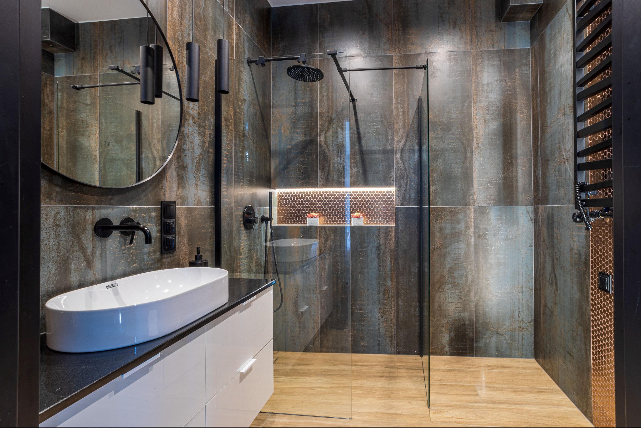 Nowoczesna łazienka z kabiną prysznicową typu walk-in, umywalką nablatową i ciemnymi płytkami imitującymi metal. Oświetlenie LED w niszy i nad lustrem.