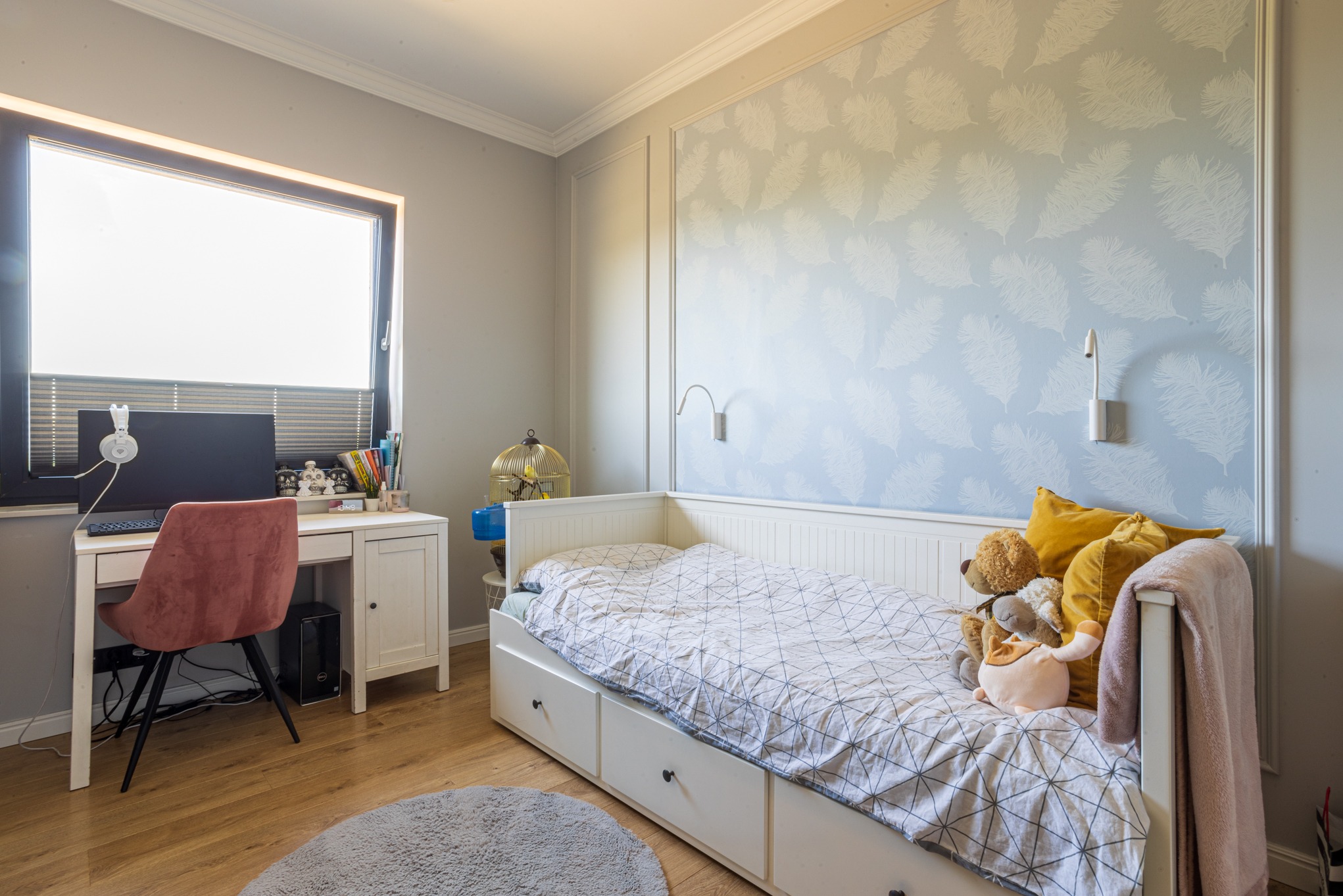 Pokój dziecięcy z łóżkiem z szufladami, biurkiem przy oknie i tapetą w pióra. Na biurku komputer i słuchawki. W pokoju drewniana podłoga i okrągły dywan.