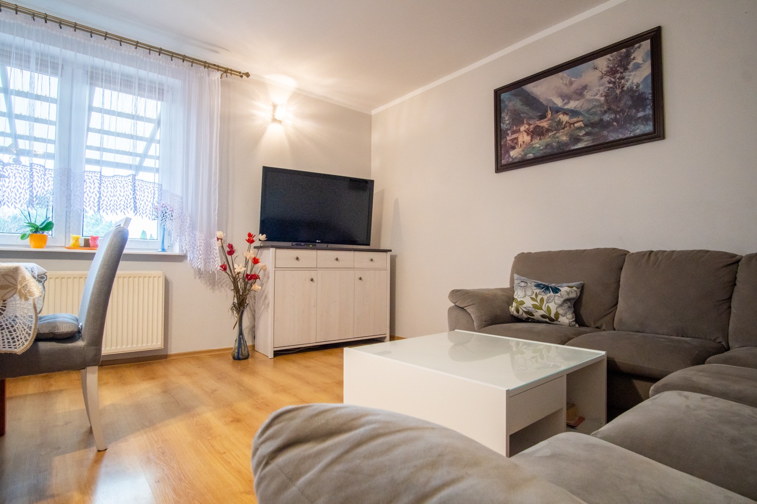 Jasne wnętrze salonu z drewnianą podłogą, szarą sofą narożną, białym stolikiem kawowym i szafką RTV pod telewizorem. Na ścianie wisi obraz z górskim pejzażem.