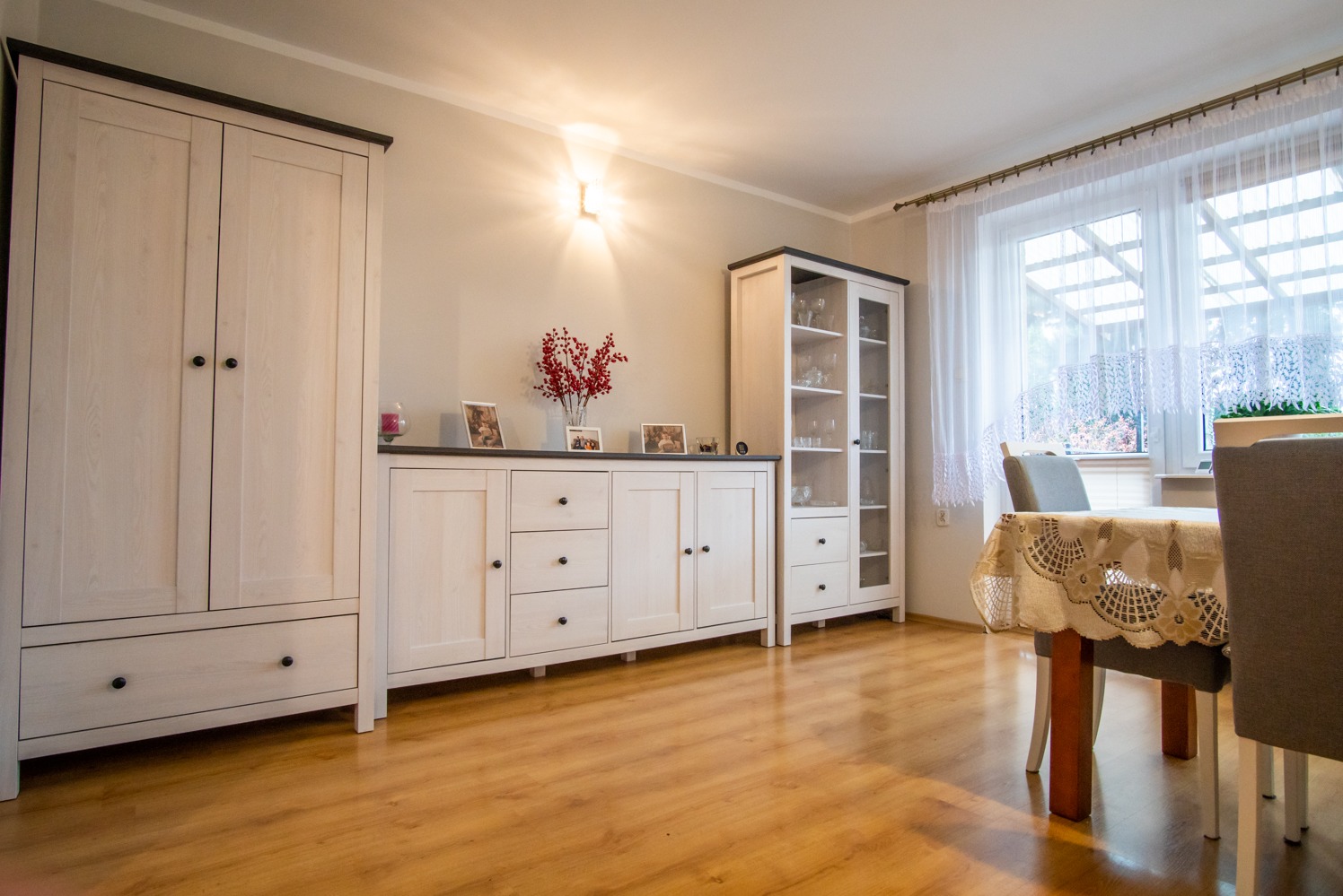 Jasne wnętrze: drewniana podłoga, białe meble z czarnymi akcentami, stół z koronkowym obrusem, okno z firankami. Przytulna aranżacja salonu.