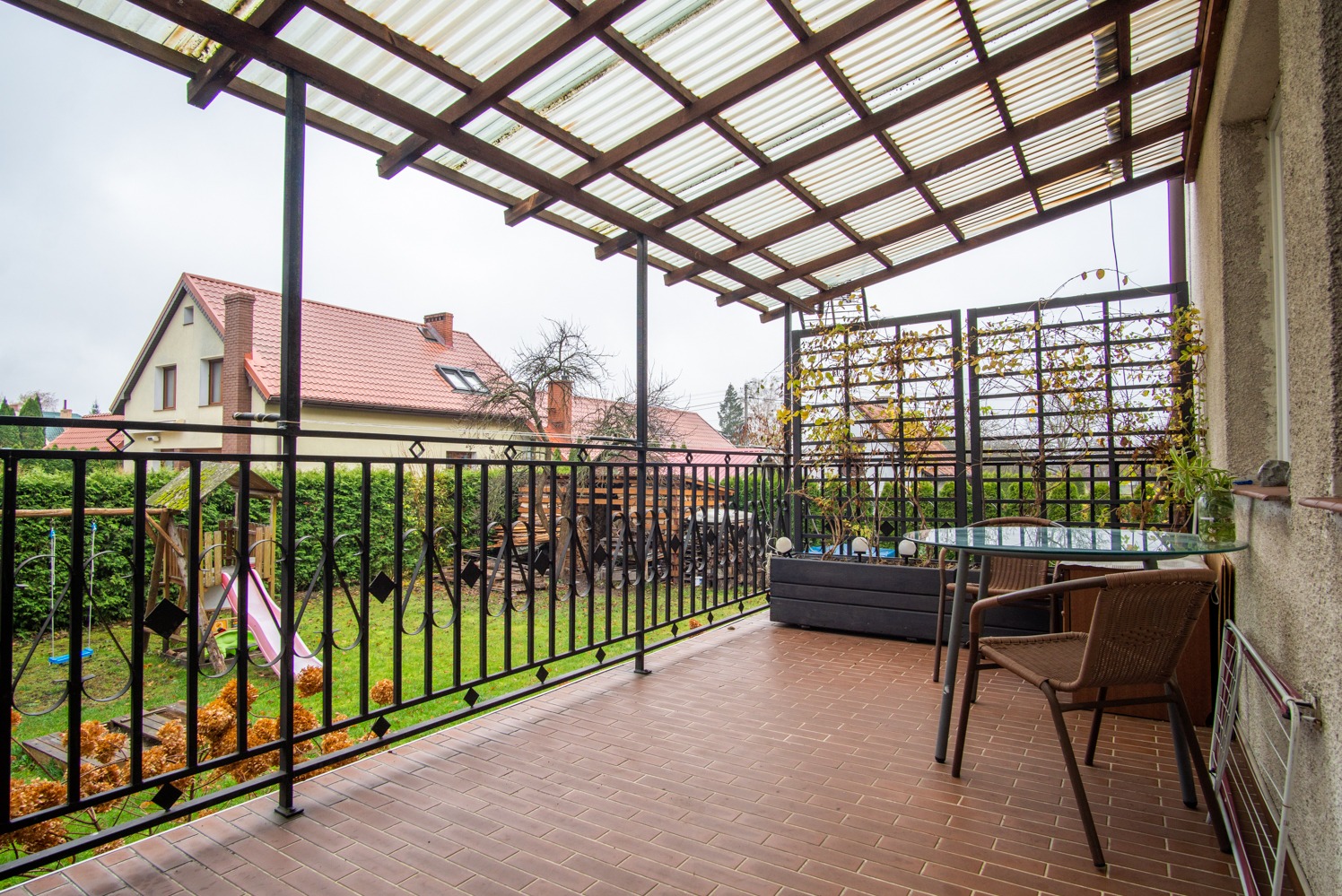 Zadaszony balkon z metalową balustradą i ceglaną podłogą. W tle dom z czerwonym dachem i zielony ogród. Na balkonie stół z krzesłami i roślina w donicy.
