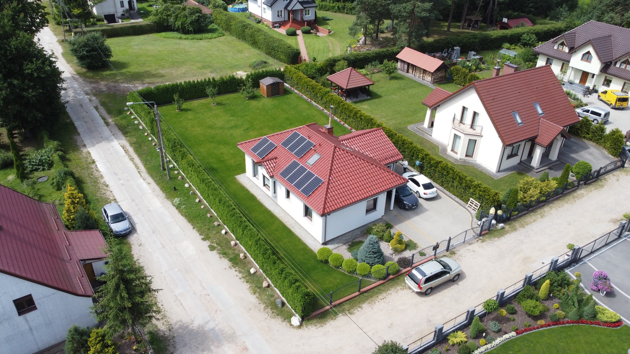 Dom jednorodzinny z panelami słonecznymi na czerwonym dachu, otoczony zielonym trawnikiem i tujami, widziany z lotu ptaka w słoneczny dzień.