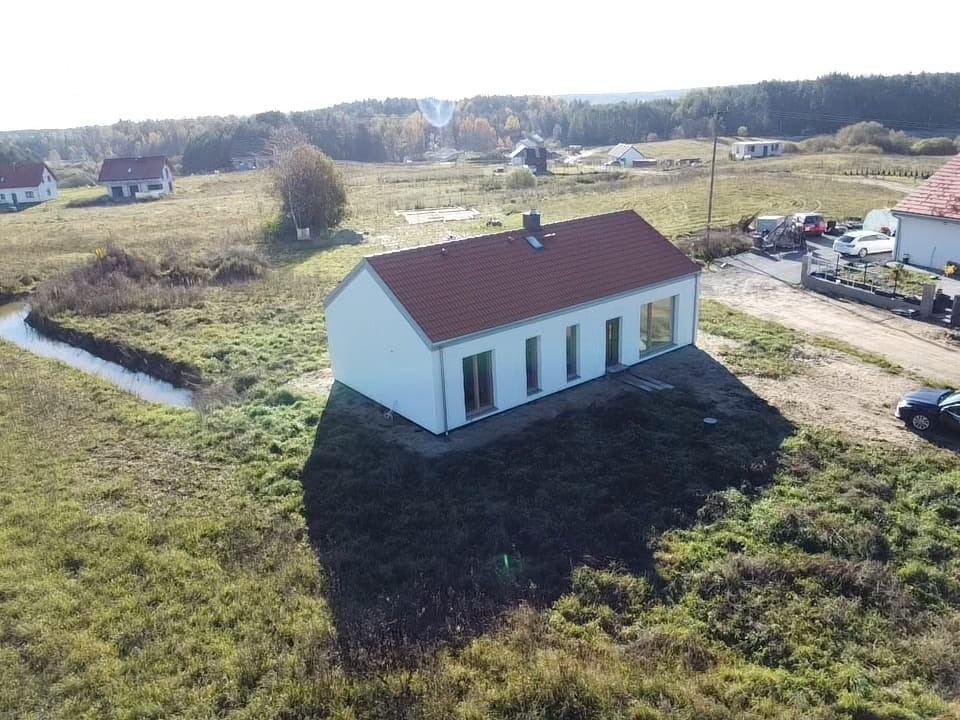 Nowoczesny, parterowy dom z czerwoną dachówką, otoczony rozległym terenem z małym ciekiem wodnym. Widok z lotu ptaka, słoneczny dzień.