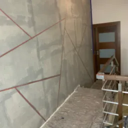Ściana w trakcie malowania z geometrycznymi wzorami wyznaczonymi czerwoną taśmą malarską, zabezpieczona podłoga i fragment schodów z drewnianą balustradą.