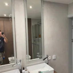 Nowoczesna łazienka po remoncie: jasne płytki, podwójna umywalka na drewnianym blacie, lustra z oświetleniem. Widoczny fragment prysznica i odbicie osoby robiącej zdjęcie.