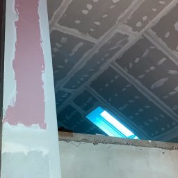 WELL DONE Bartłomiej Paliga - Sufit z płyt gipsowo-kartonowych w trakcie szpachlowania, z widocznym oknem dachowym i fragmentem ściany pokrytej gładzią. Widoczny filar z różowym podkładem.