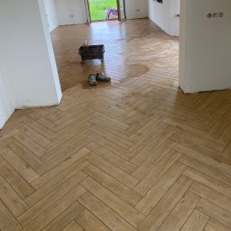 WELL DONE Bartłomiej Paliga - Ułożona parkietowa podłoga z płytek w jodełkę, wiadro glazurnicze na kółkach i robocze buty w pomieszczeniu w trakcie prac wykończeniowych.