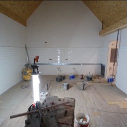 WELL DONE Bartłomiej Paliga - Wnętrze w trakcie prac glazurniczych: białe, prostokątne płytki ułożone na ścianach, narzędzia i materiały rozłożone na podłodze. Widoczna lampa budowlana i fragment drewnianej konstrukcji dachu.