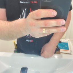 WELL DONE Bartłomiej Paliga - Odbicie w lustrze: glazurnik w koszulce Sopro robi zdjęcie telefonem, widoczna umywalka i narzędzia w pasie. Ujęcie z bliska na dłoń z obrączką.