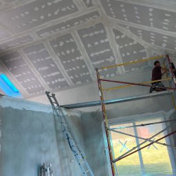 WELL DONE Bartłomiej Paliga - Skośny sufit poddasza po położeniu płyt gips-karton z widocznymi łączeniami, pracownik na rusztowaniu wygładza powierzchnię. Okna dachowe.