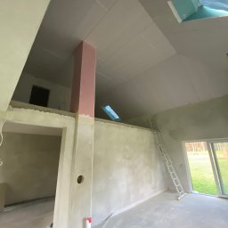 WELL DONE Bartłomiej Paliga - Surowe wnętrze z płytami gipsowo-kartonowymi na skośnym suficie, drabina oparta o ścianę, widok na zewnątrz przez okno. W tle antresola.