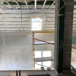 WELL DONE Bartłomiej Paliga - Poddasze z ociepleniem pianą PUR, metalowa konstrukcja sufitu, rusztowanie przy ścianie. Widoczne narzędzia i materiały budowlane. Prace wykończeniowe w toku.