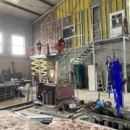 Montaż izolacji wewnątrz hali: pracownicy na podnośniku układają wełnę mineralną w metalową konstrukcję ściany, widoczne materiały i narzędzia.