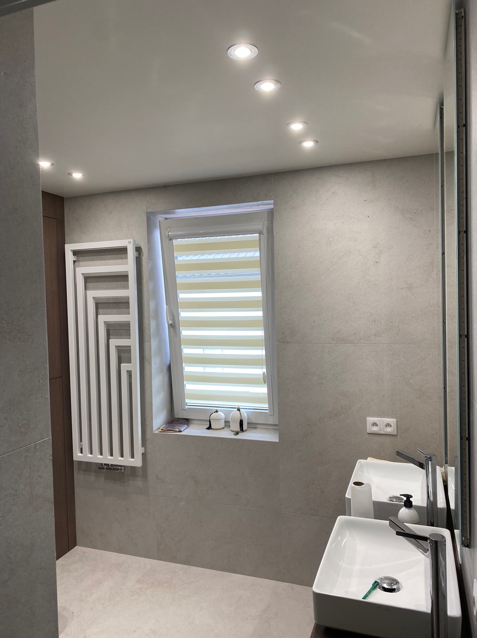 Jasna łazienka z dwoma umywalkami, grzejnikiem dekoracyjnym i oknem z żaluzjami poziomymi. Minimalistyczny design, płytki w odcieniach szarości, oświetlenie punktowe.