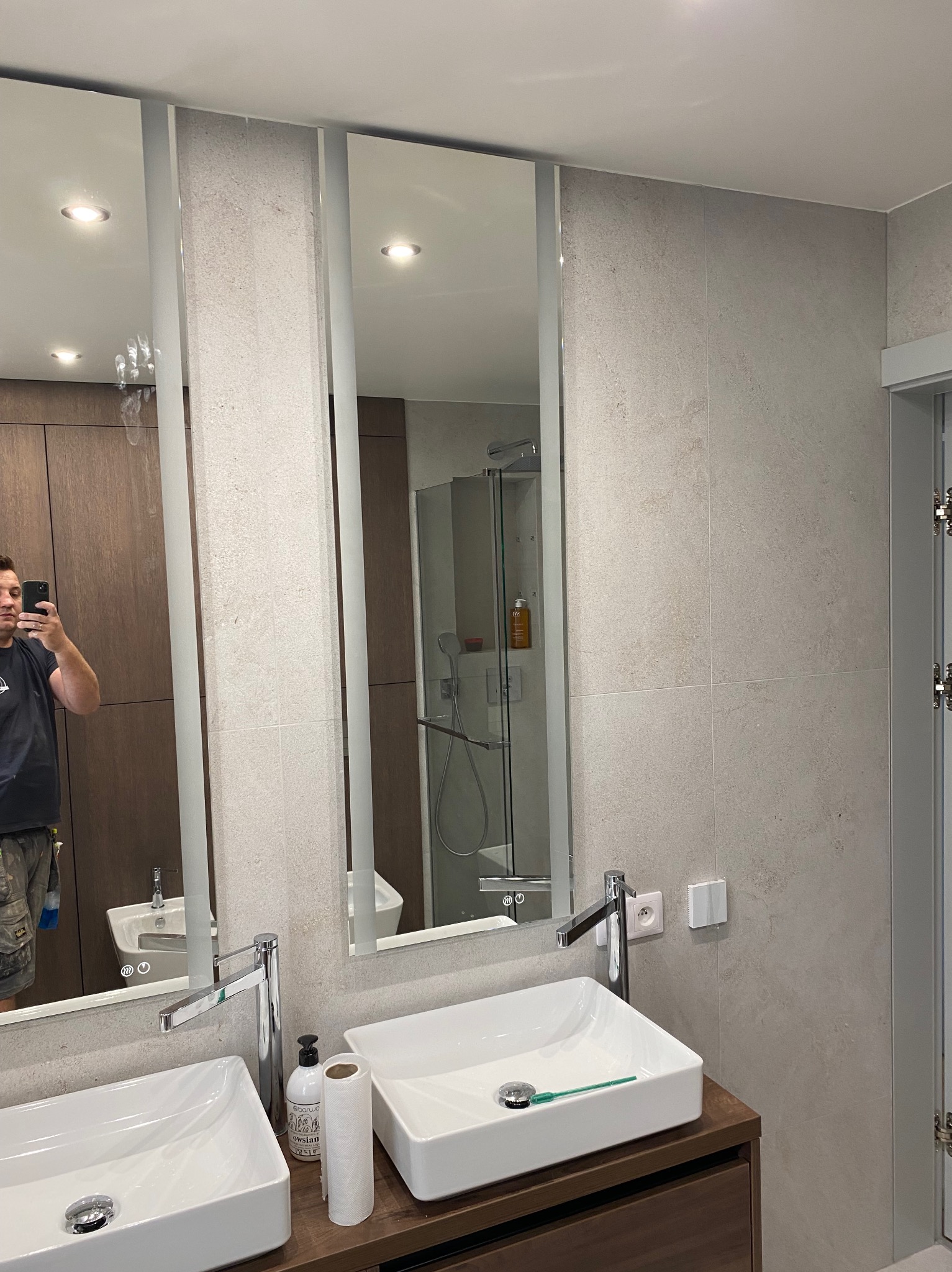 Nowoczesna łazienka po remoncie: jasne płytki, podwójna umywalka na drewnianym blacie, lustra z oświetleniem. Widoczny fragment prysznica i odbicie osoby robiącej zdjęcie.