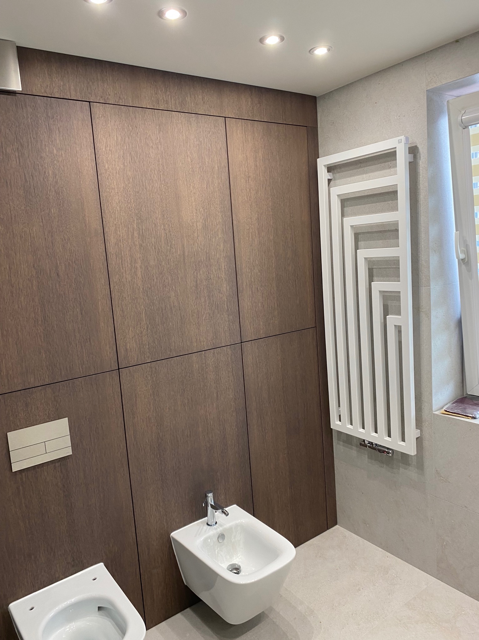 Nowoczesna łazienka z drewnianą zabudową, białym bidetem i grzejnikiem drabinkowym. Minimalistyczny design, jasne płytki i punktowe oświetlenie.