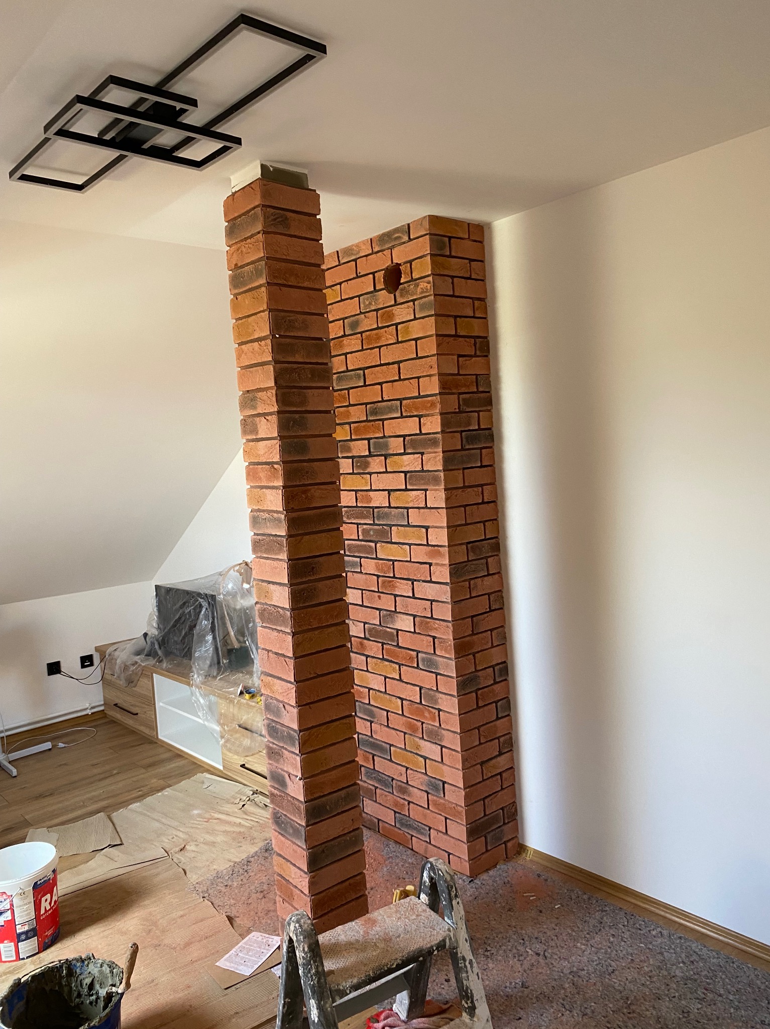 Wnętrze poddasza z elementami dekoracyjnymi z cegły klinkierowej. Widoczne narzędzia i materiały budowlane, co sugeruje prace wykończeniowe w trakcie realizacji.