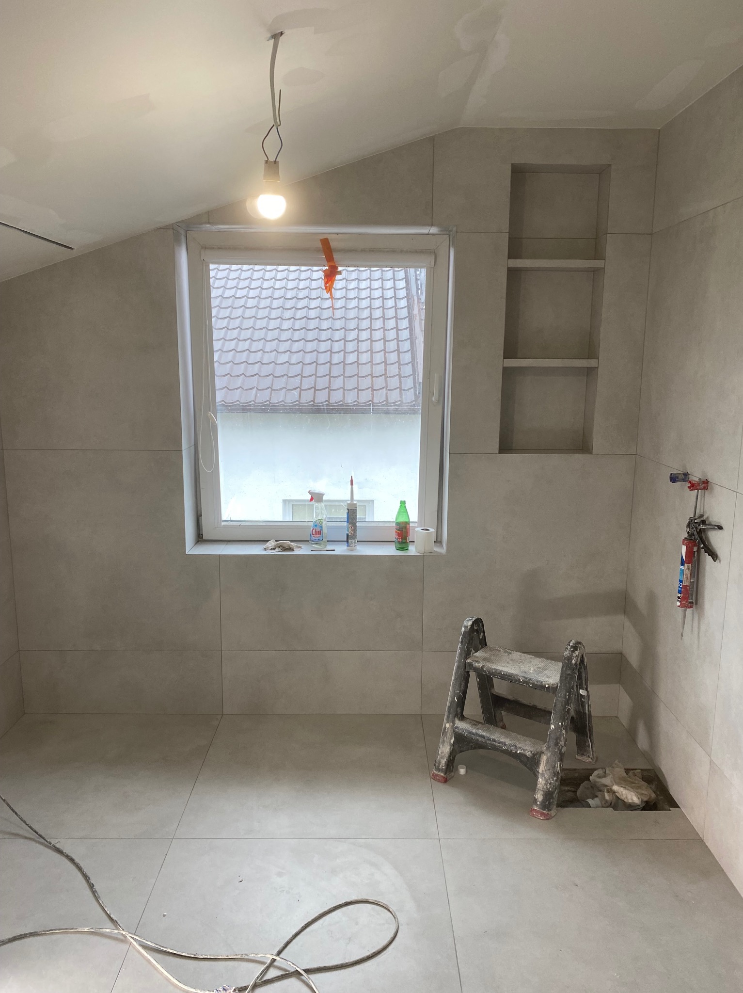 Wykończona łazienka na poddaszu z oknem i wnękami, wyłożona jasnymi płytkami. Widoczna drabina i narzędzia wskazują na etap prac wykończeniowych.