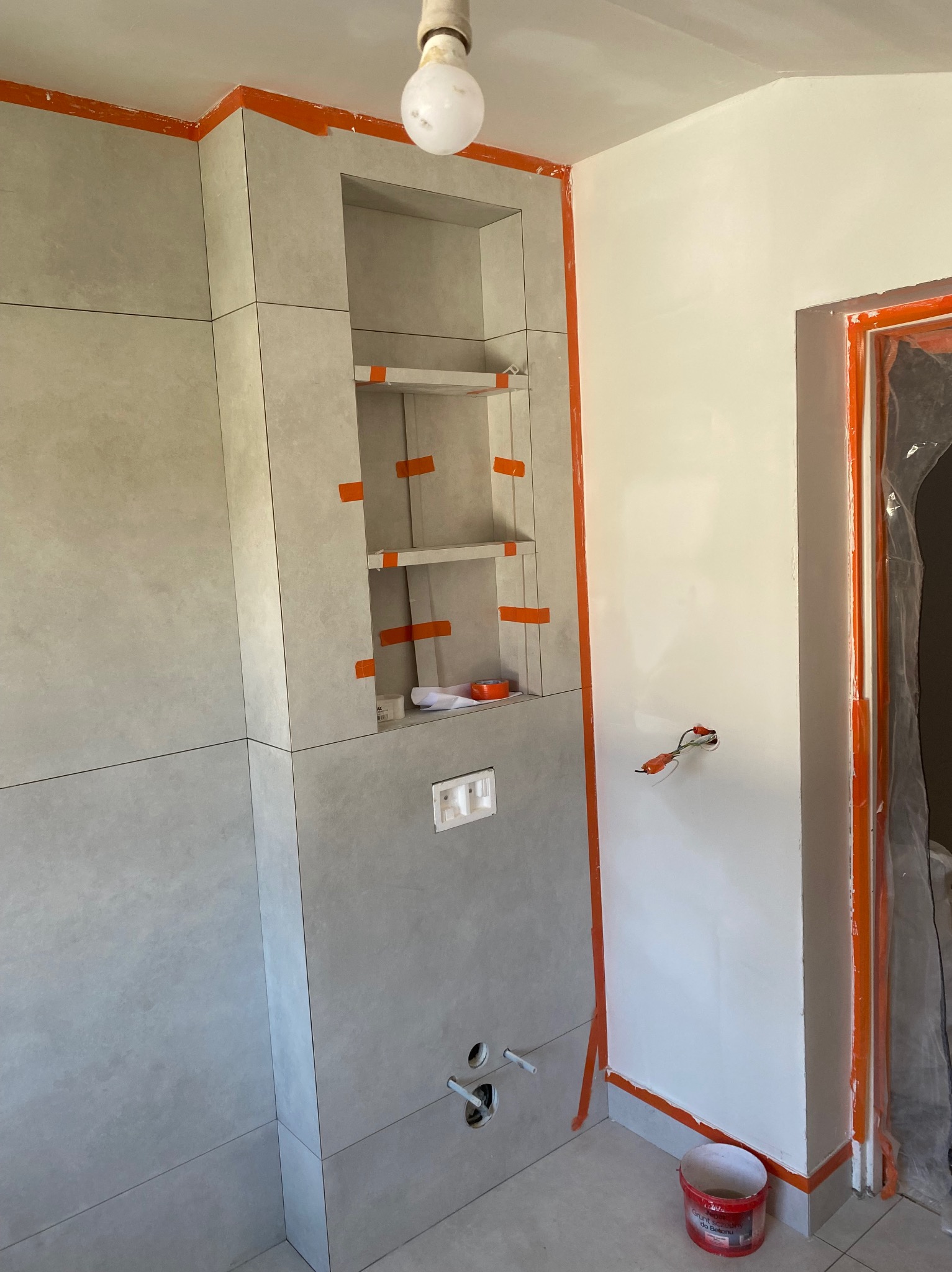 Świeżo ułożone, duże, matowe płytki w łazience z wnęką, zabezpieczone pomarańczową taśmą malarską. Widoczne przyłącza wodne i puszka elektryczna.