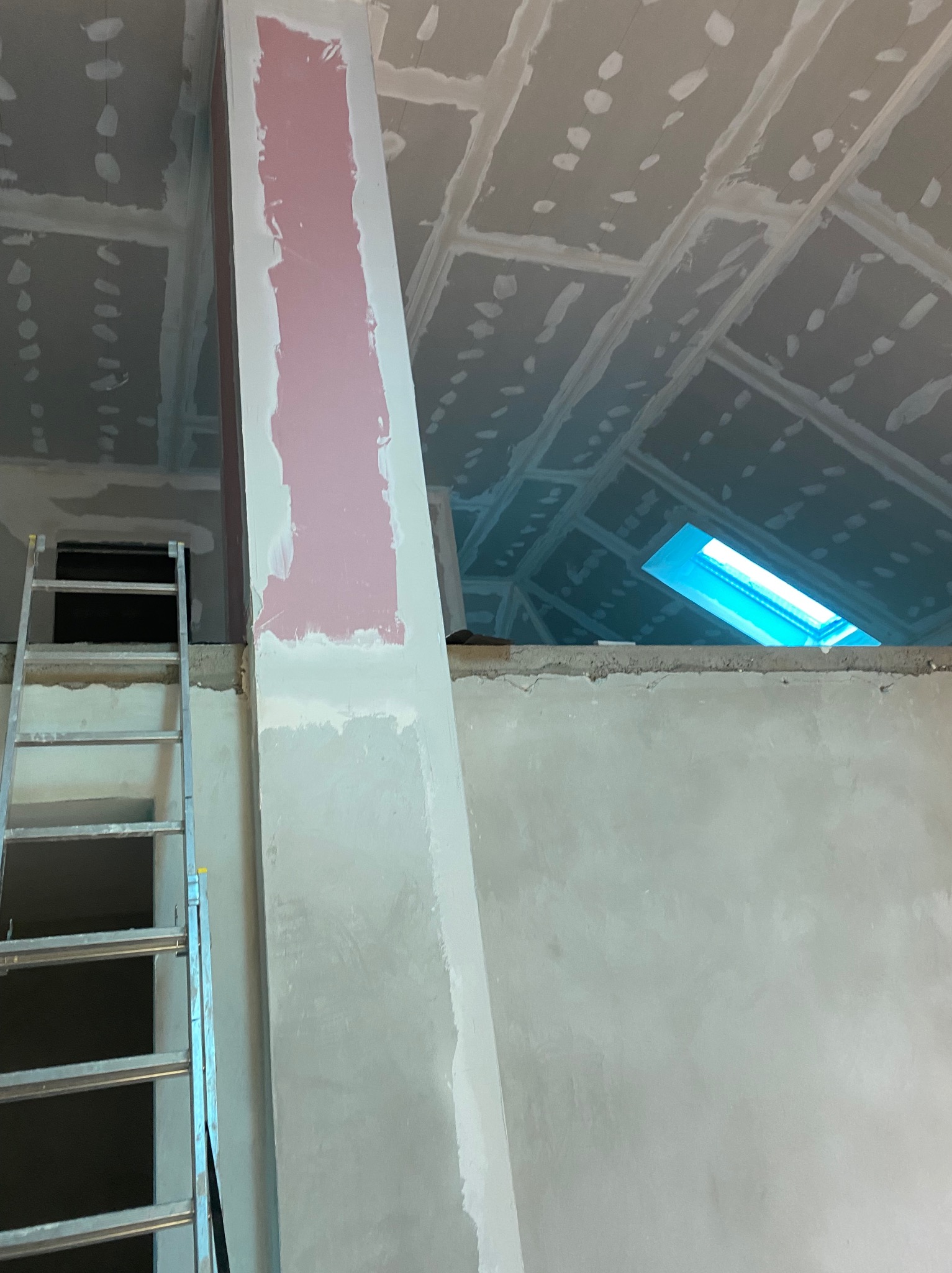 Poddasze w trakcie wykańczania: płyty gipsowe na skosach, szpachlowana kolumna, drabina przystawiona do ściany. Widoczne okno dachowe.