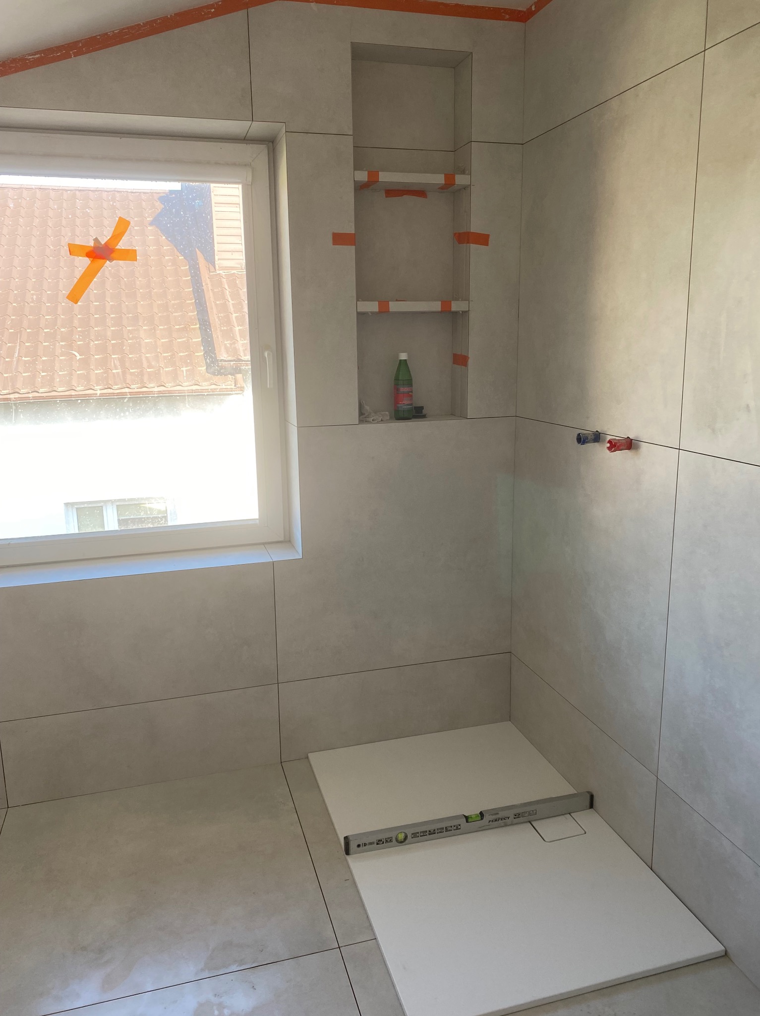 Łazienka w trakcie prac: jasne płytki, wnęka z półkami zabezpieczona taśmą, poziomica na brodziku. Widok na okno z zewnątrz.