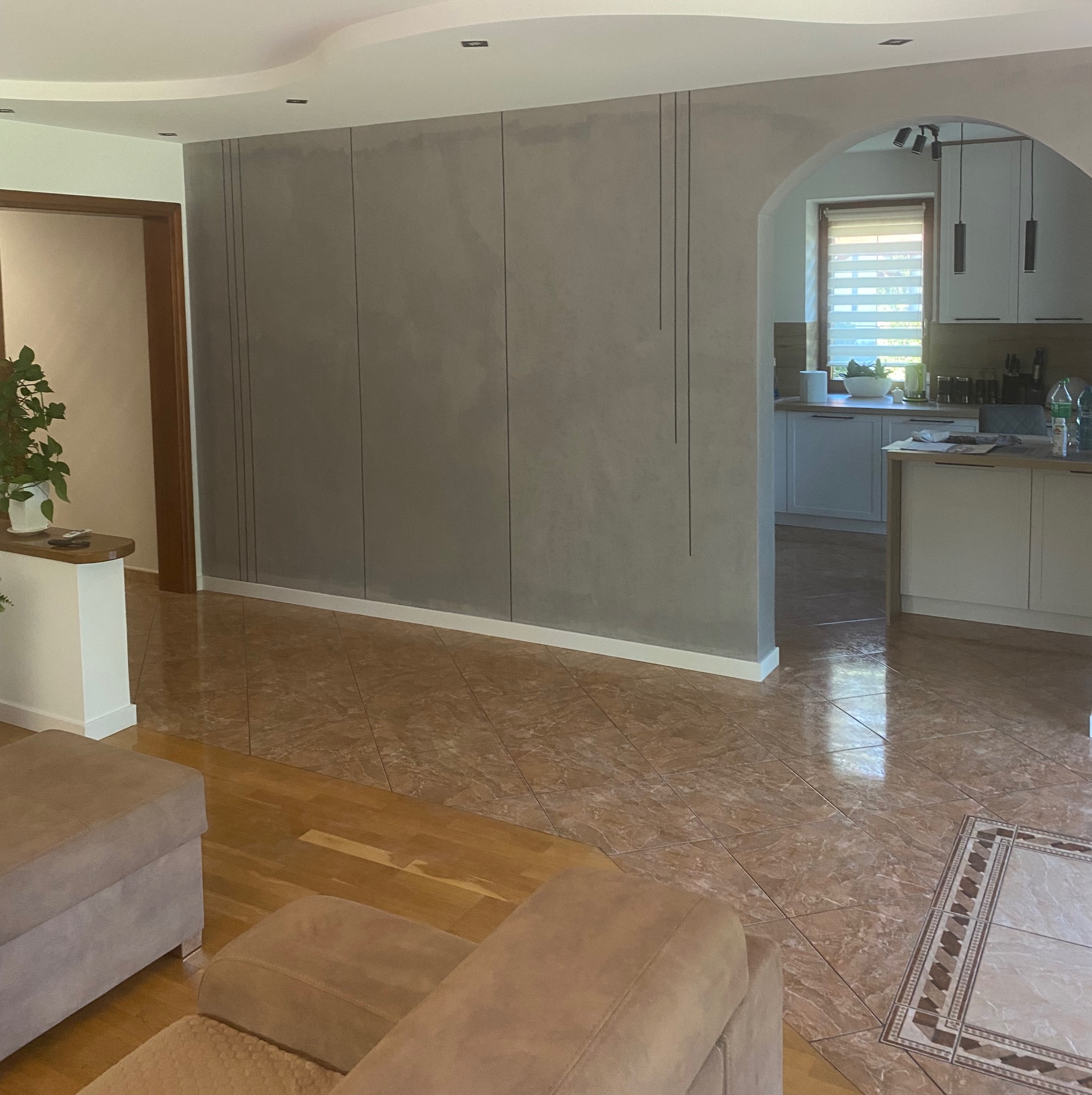 Wykończone wnętrze salonu z dekoracyjną ścianą w odcieniach szarości, z elementami pionowych linii. Widoczna kuchnia w tle i fragment podłogi z płytek.