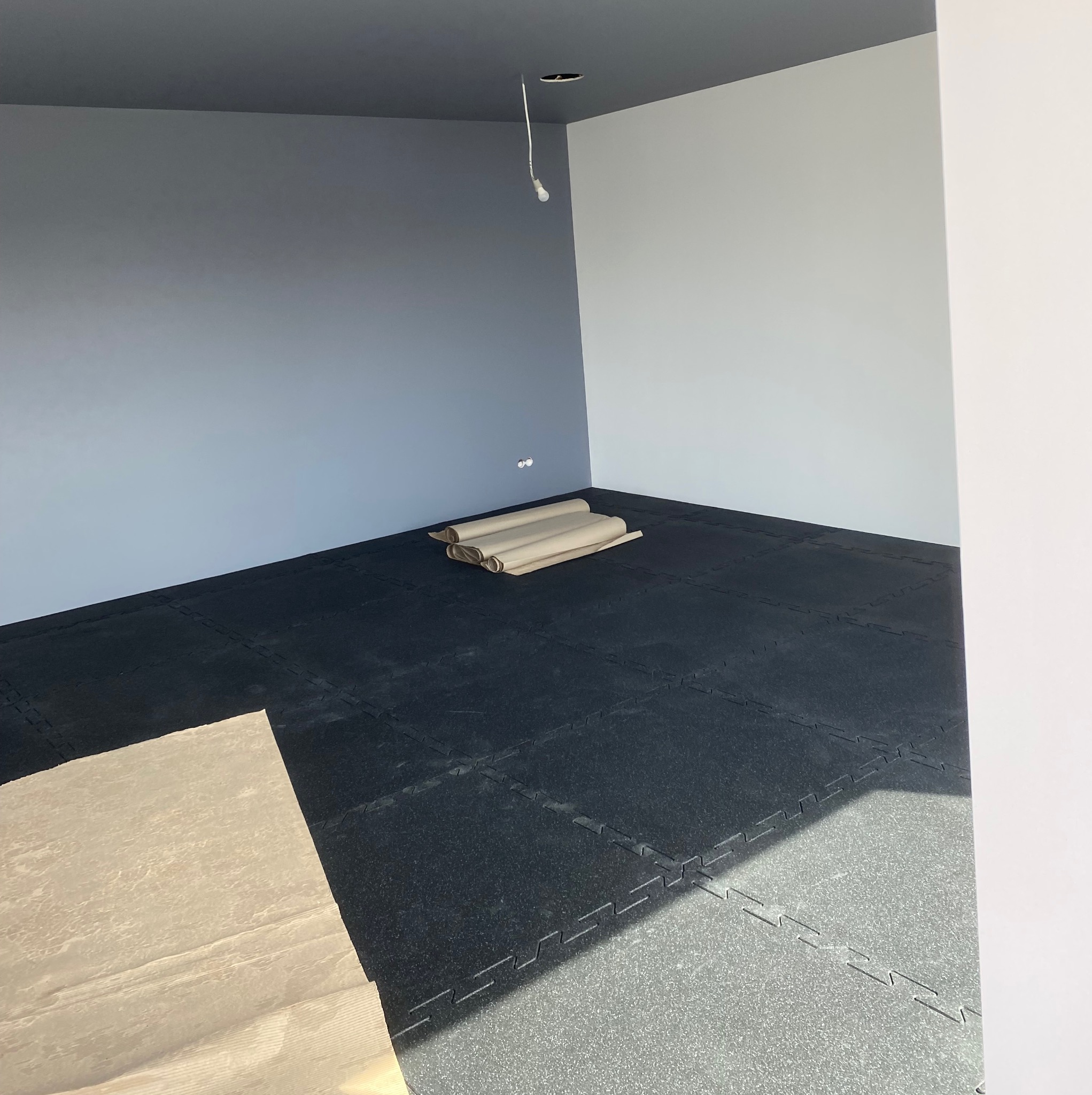 Pokój po montażu czarnej podłogi z gumowych puzzli. Na podłodze leżą zwinięte arkusze papieru ochronnego, ściany pomalowane na biało i szaro.