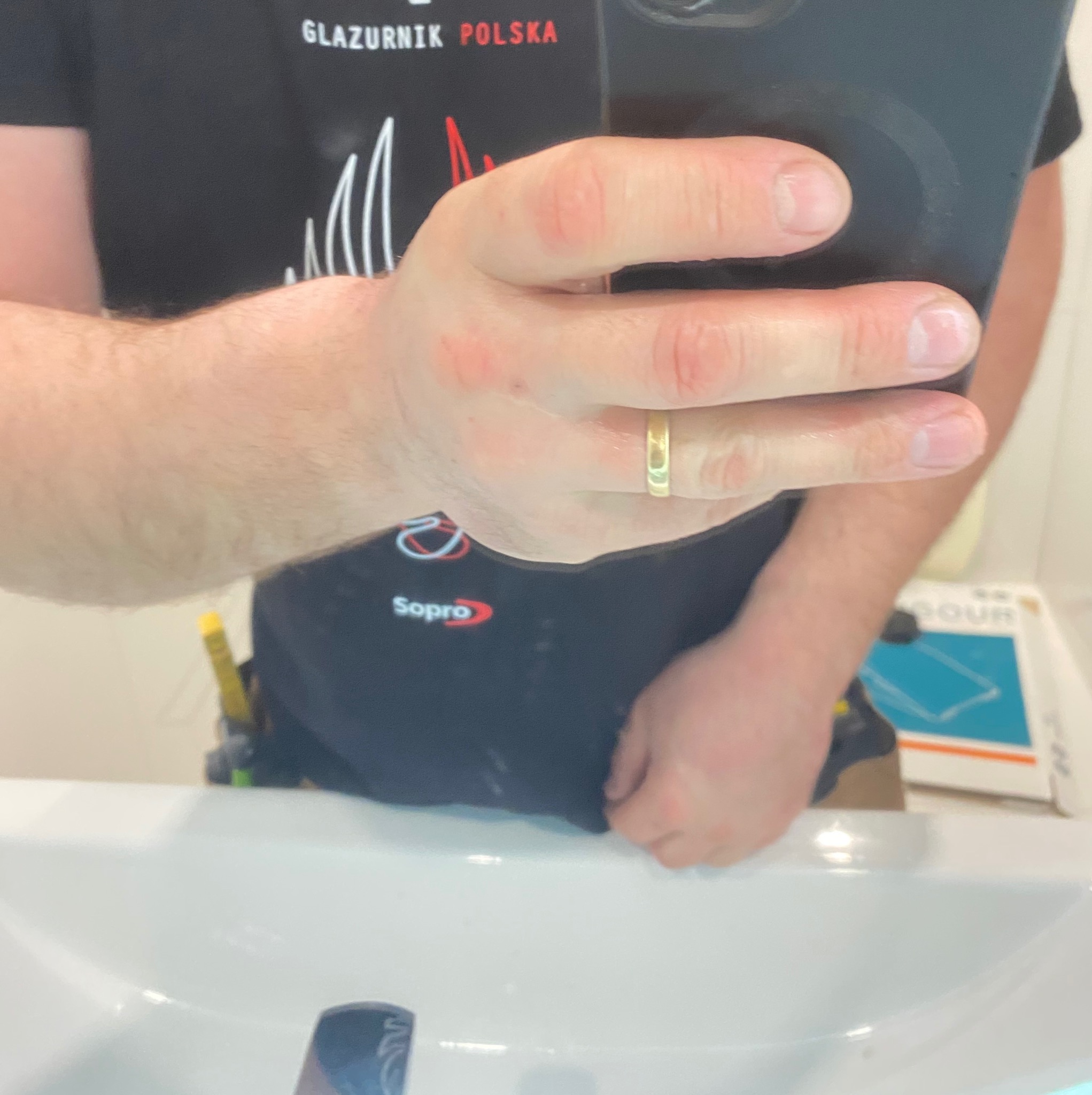 Odbicie w lustrze: glazurnik w koszulce Sopro robi zdjęcie telefonem, widoczna umywalka i narzędzia w pasie. Ujęcie z bliska na dłoń z obrączką.