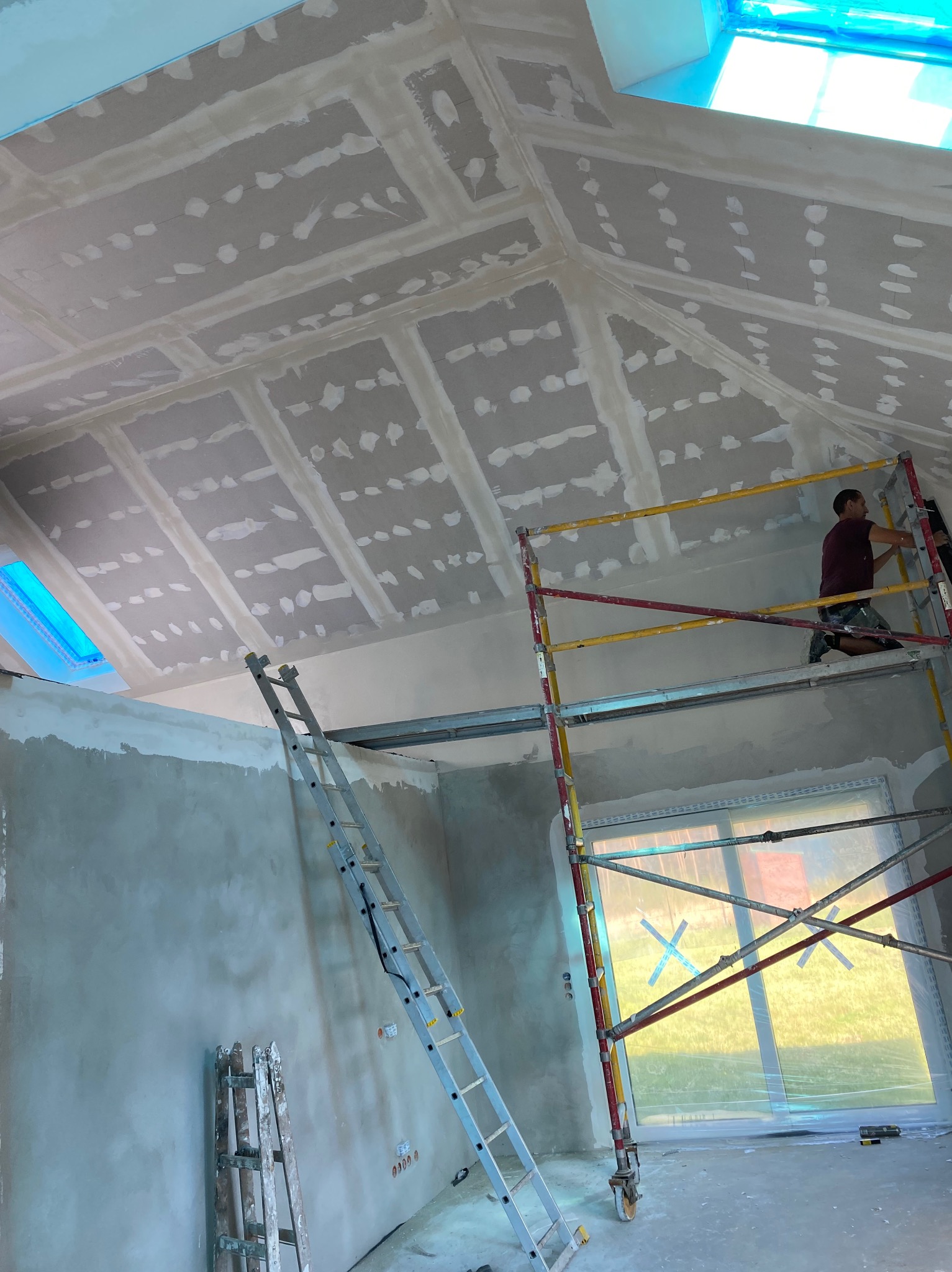 Skośny sufit poddasza po położeniu płyt gips-karton z widocznymi łączeniami, pracownik na rusztowaniu wygładza powierzchnię. Okna dachowe.