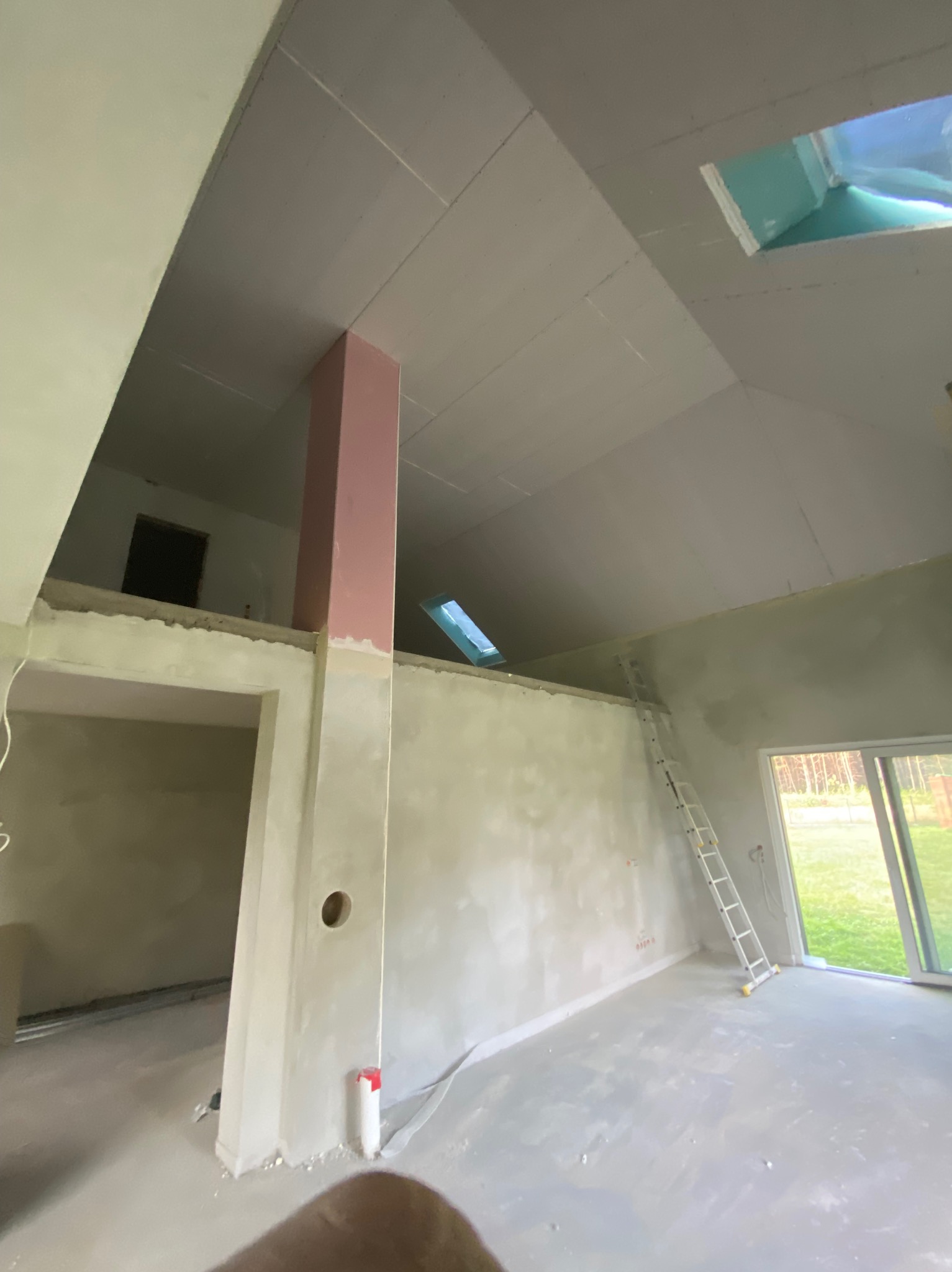 Surowe wnętrze z płytami gipsowo-kartonowymi na skośnym suficie, drabina oparta o ścianę, widok na zewnątrz przez okno. W tle antresola.