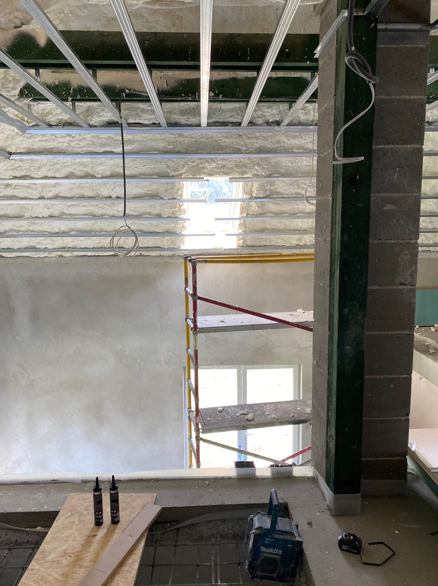 Poddasze z ociepleniem pianą PUR, metalowa konstrukcja sufitu, rusztowanie przy ścianie. Widoczne narzędzia i materiały budowlane. Prace wykończeniowe w toku.