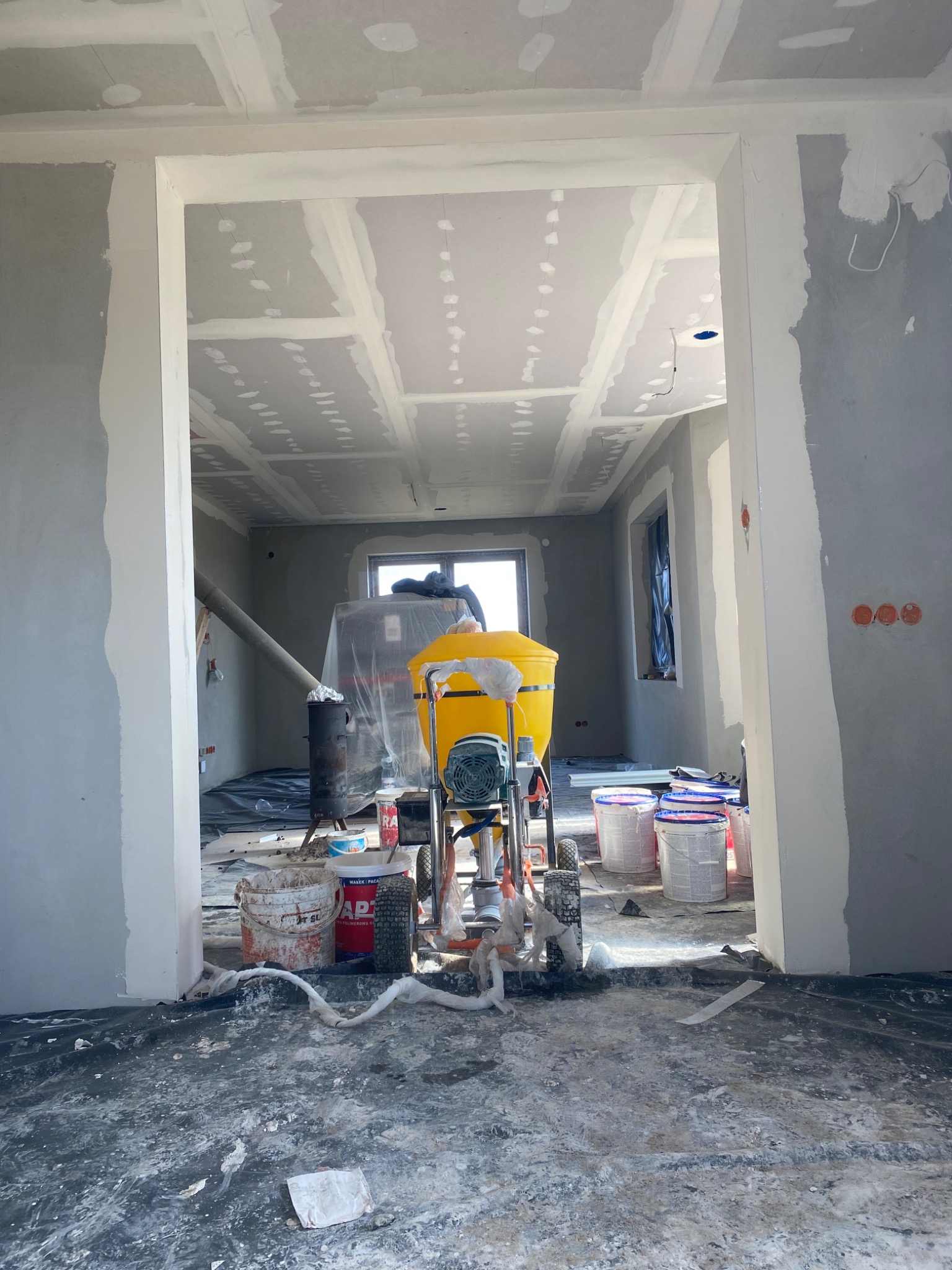 Wnętrze w trakcie prac wykończeniowych: ściany i sufit po szpachlowaniu, widoczna maszyna do gładzi, podłoga zabezpieczona folią. Prace przygotowawcze przed malowaniem.