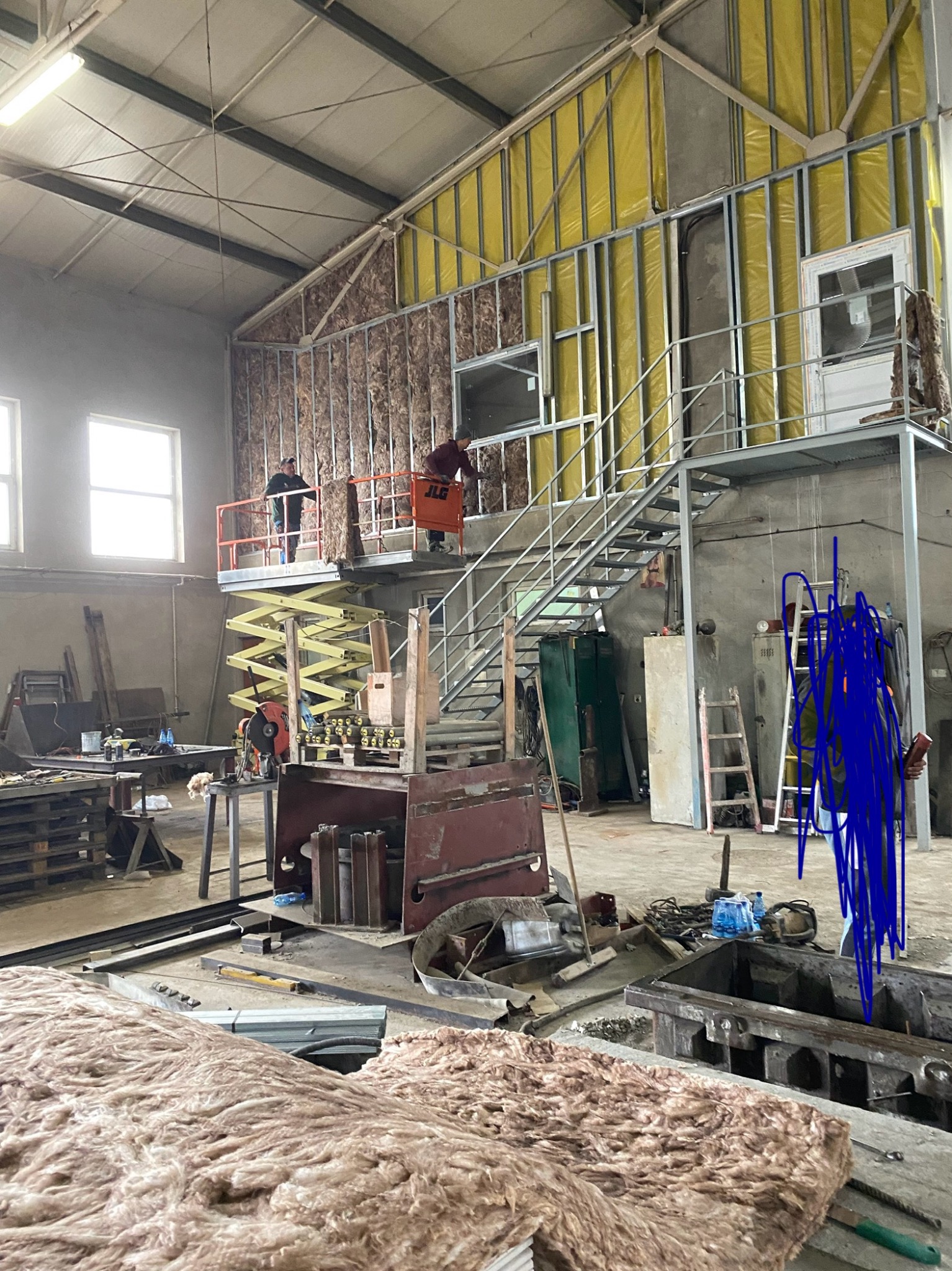 Montaż izolacji wewnątrz hali: pracownicy na podnośniku układają wełnę mineralną w metalową konstrukcję ściany, widoczne materiały i narzędzia.