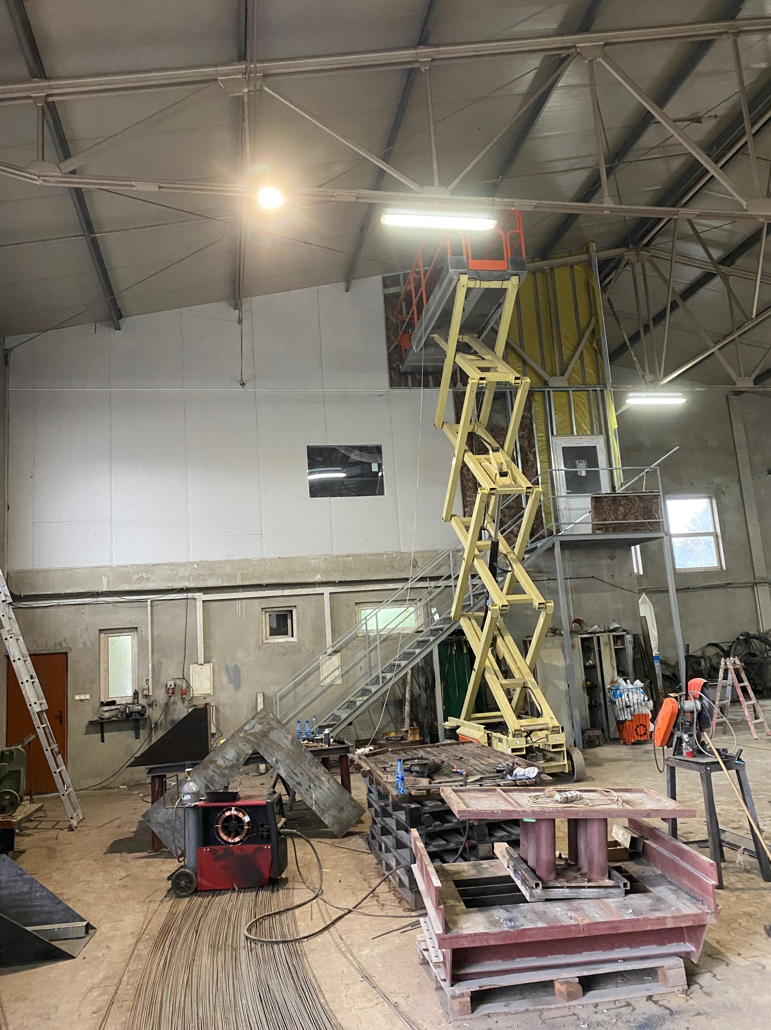 Wnętrze hali w trakcie prac. Widoczna podnośnik nożycowy, narzędzia, materiały budowlane i konstrukcja metalowa. Ściany w surowym stanie, widoczne płyty gipsowe.