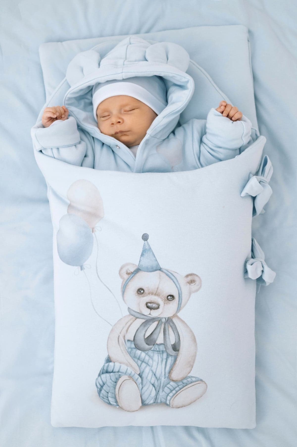 Śpiący noworodek w błękitnym kombinezonie z kapturem i czapeczce, otulony w błękitny kocyk z rysunkiem misia w czapeczce urodzinowej i balonami.