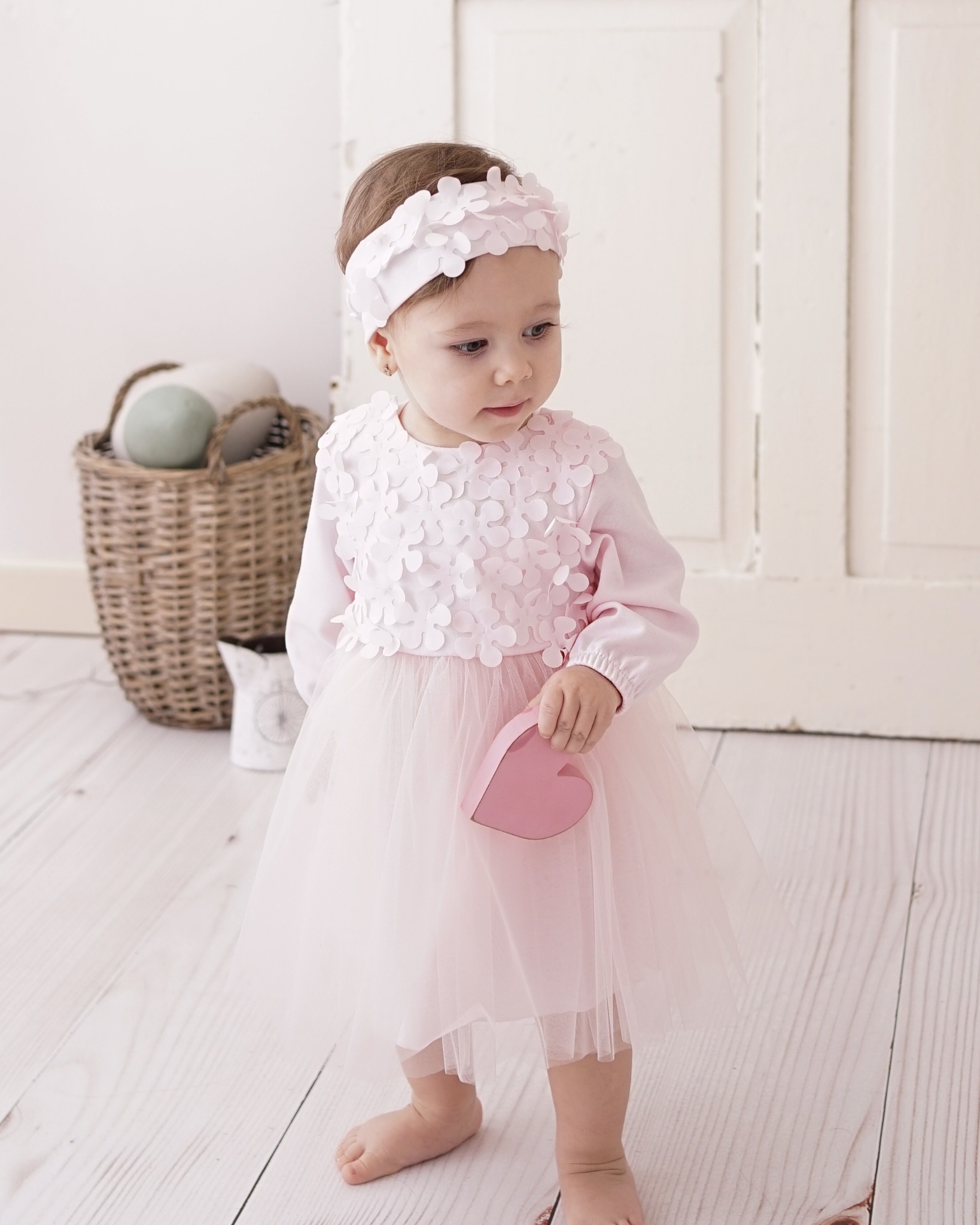 Urocza dziewczynka w różowej sukience z tiulu i opasce z kwiatami, trzymająca pudełko w kształcie serca, pozuje na jasnej drewnianej podłodze.