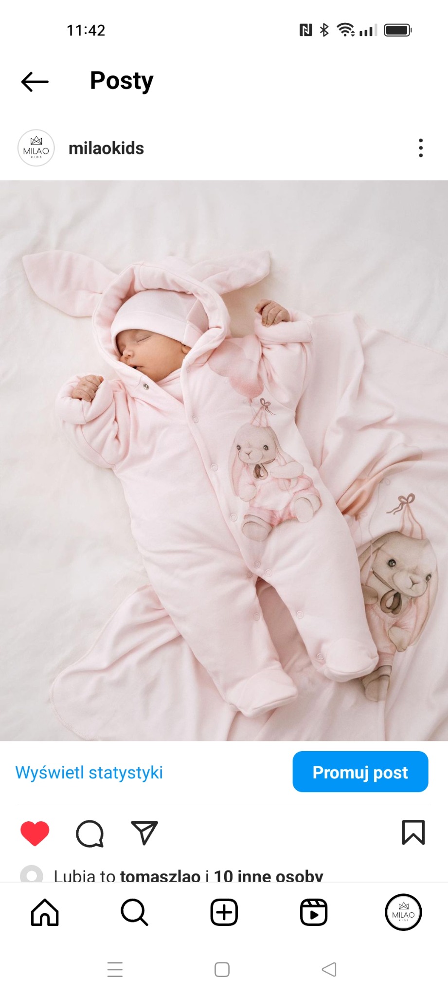 Śpiące niemowlę w różowym kombinezonie z kapturem w kształcie króliczych uszu, leżące na kocyku z motywem króliczków.